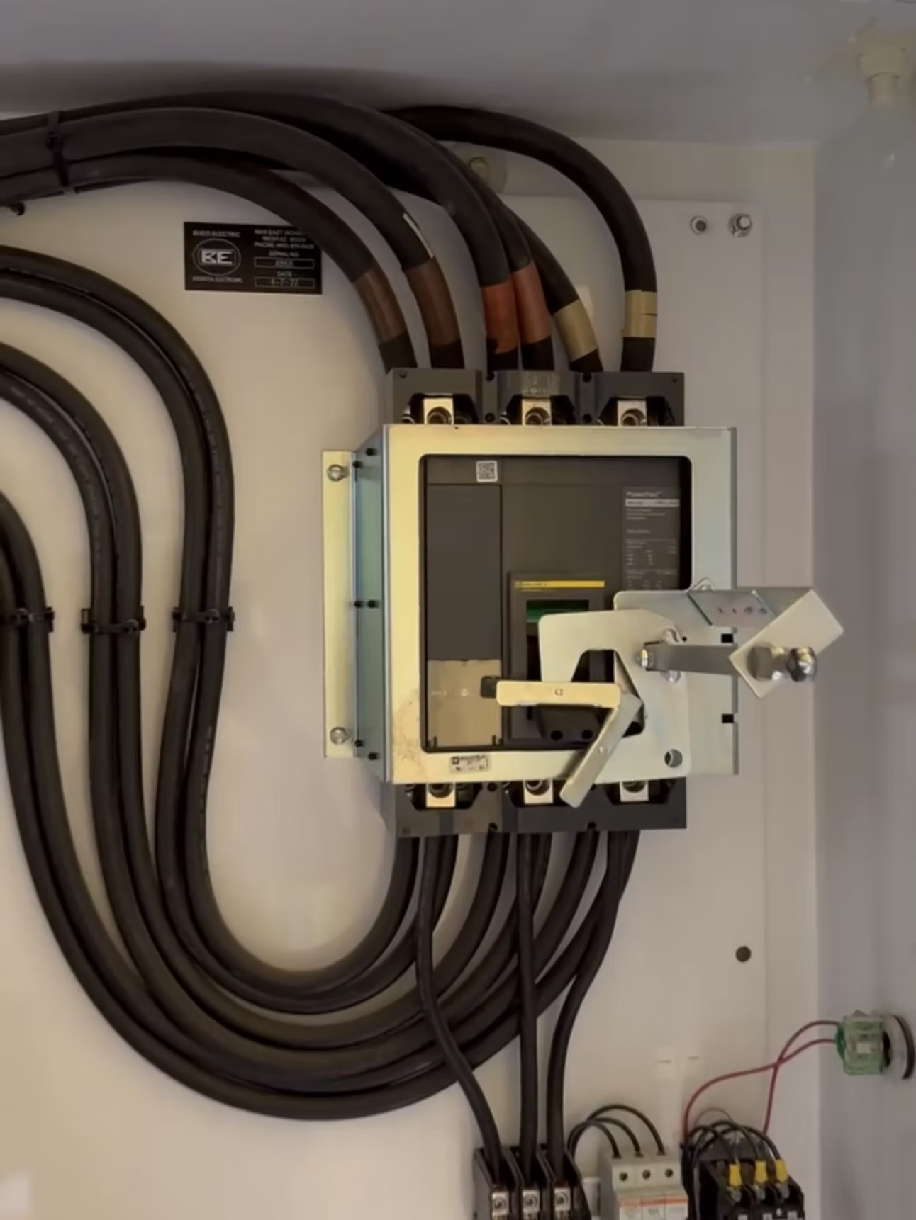 Electrical panel with large black cables connected to a circuit breaker with a silver handle, inside a white cabinet.
