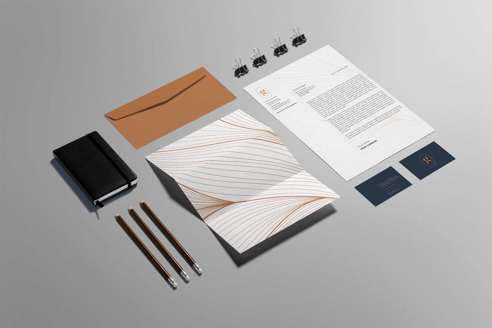 Material de oficina dispuesto de forma plana con cuaderno, bolígrafos, sobres, tarjetas de visita y hojas impresas sobre un fondo gris.