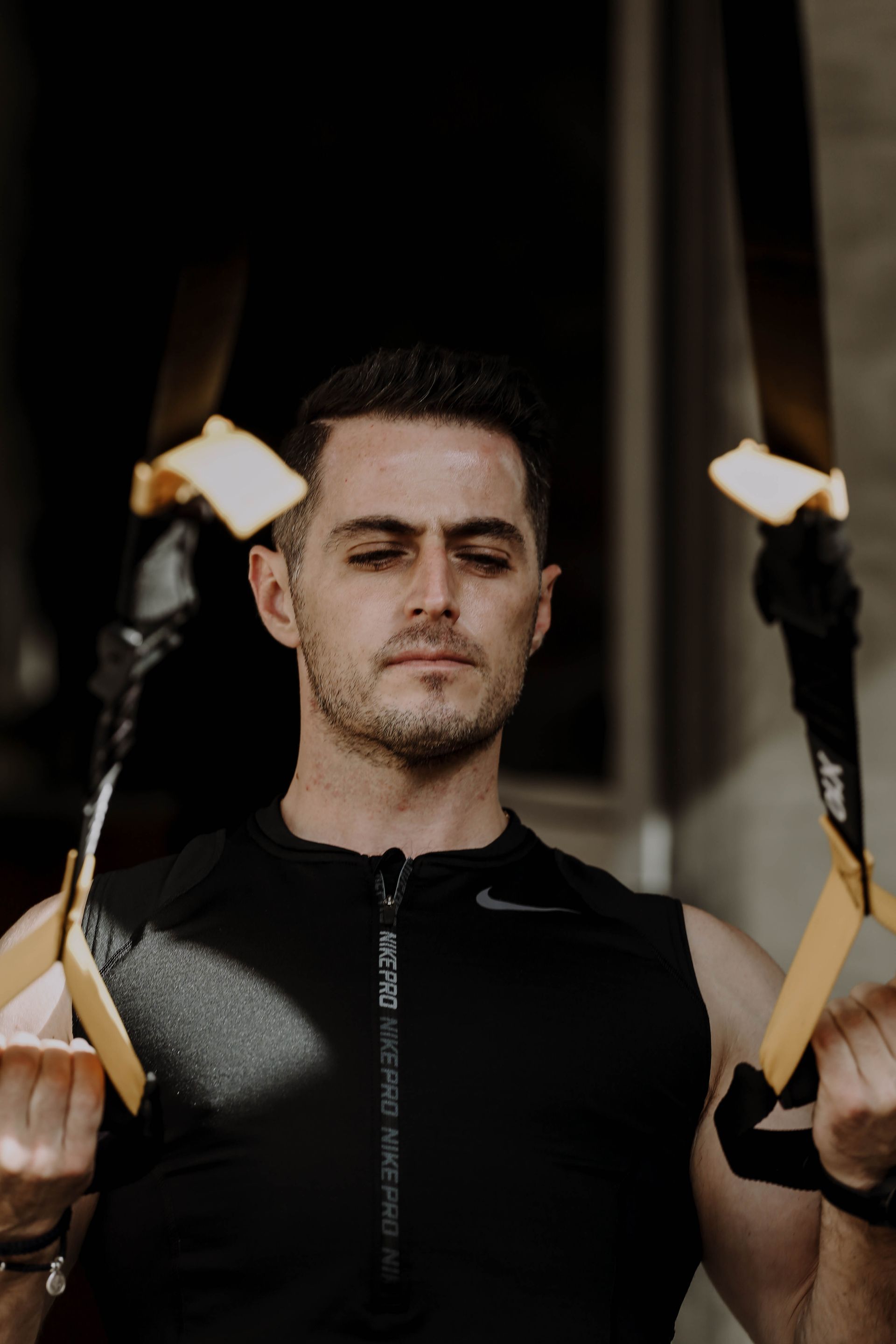 Hombre con ropa deportiva negra sosteniendo correas de suspensión, con los ojos cerrados, en un gimnasio oscuro.