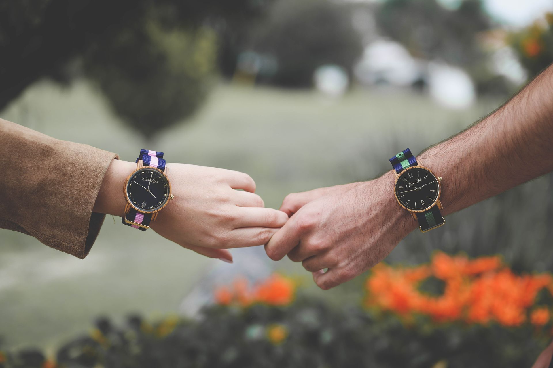 Dos manos tocándose, cada una con un reloj, con flores borrosas al fondo.