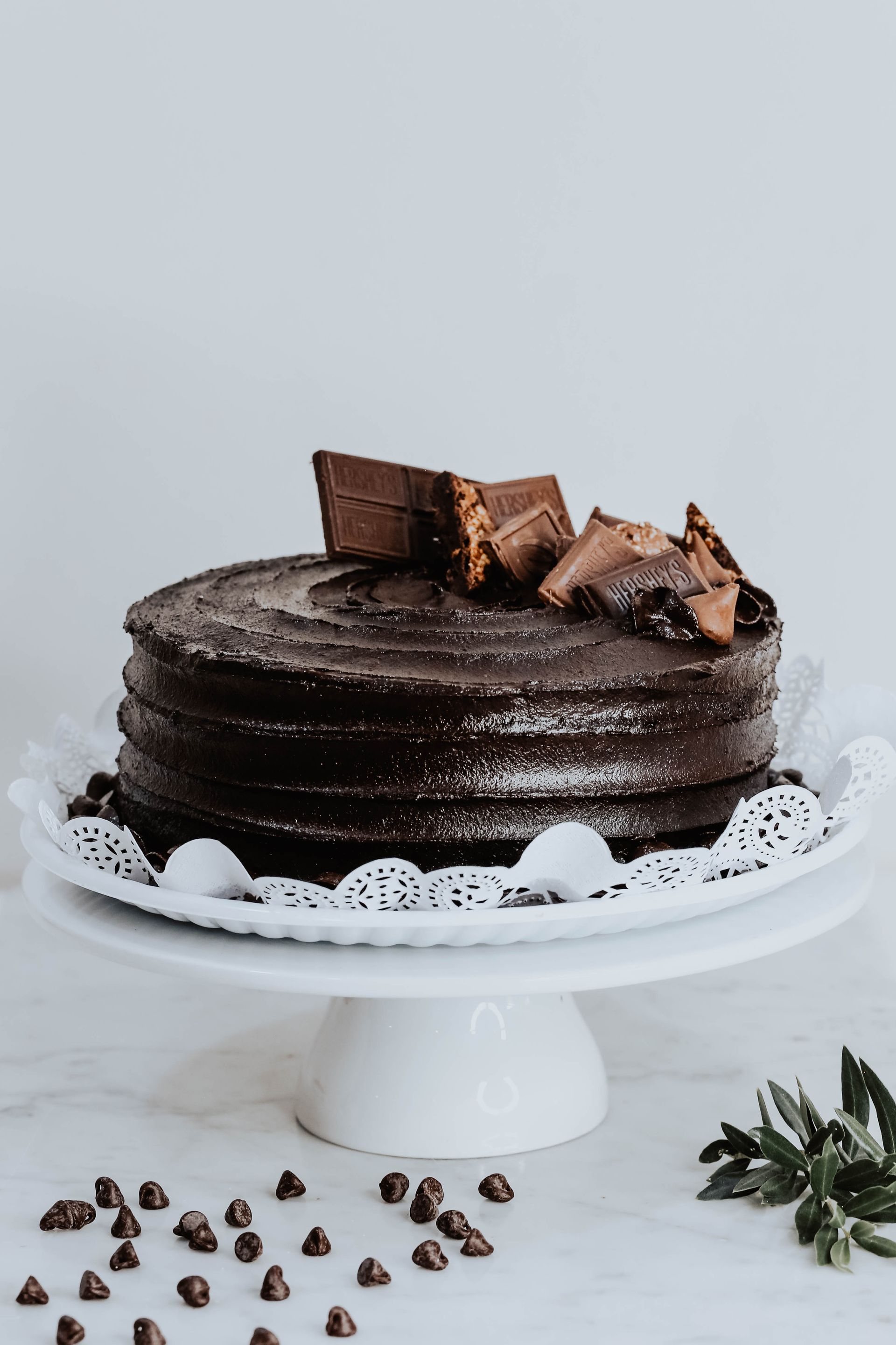 Pastel de chocolate en capas sobre una base blanca, cubierto con trozos de chocolate y rodeado de chispas de chocolate.