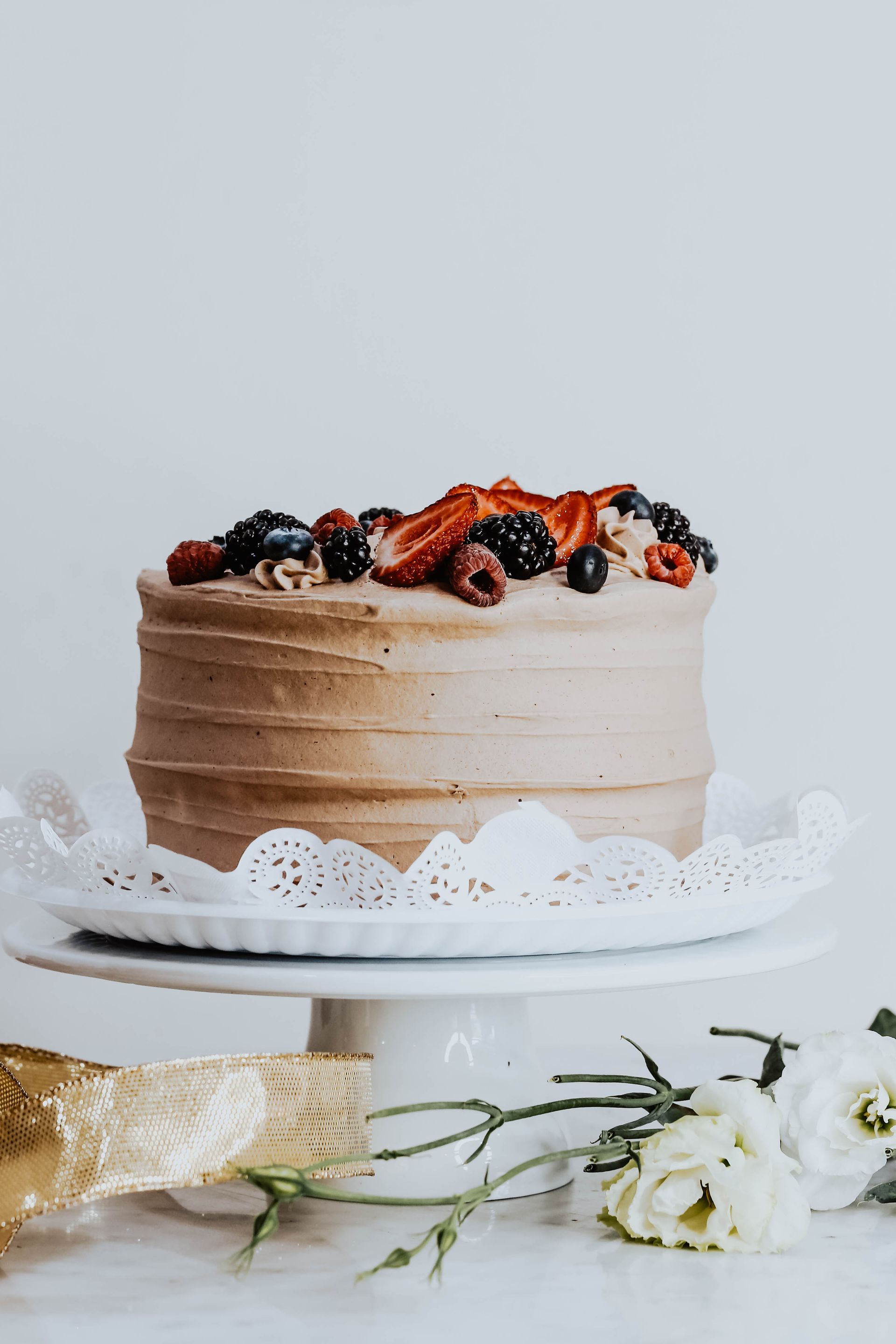 Pastel de chocolate cubierto con frutos rojos en un plato blanco, con flores sobre un fondo claro.