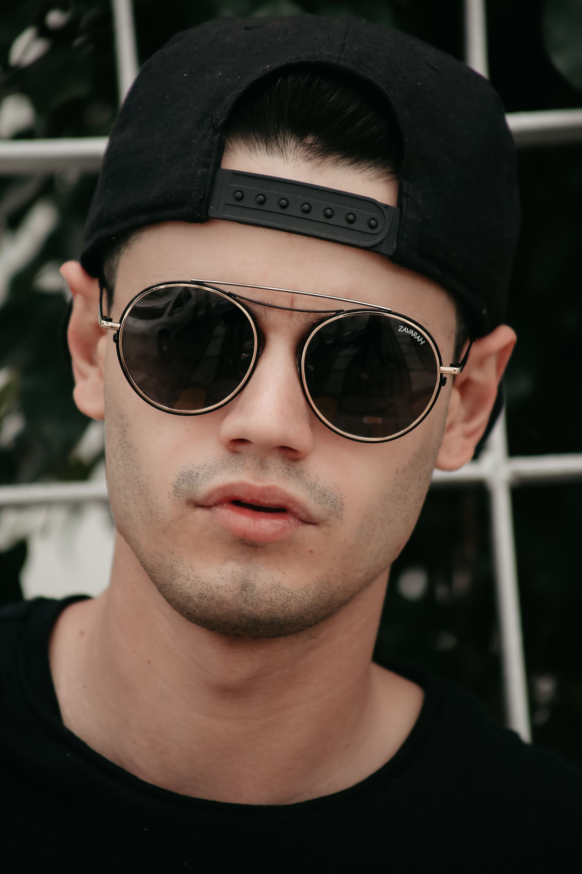 Retrato de una persona con gorra negra, gafas de sol redondas y camisa negra, sobre un fondo oscuro.
