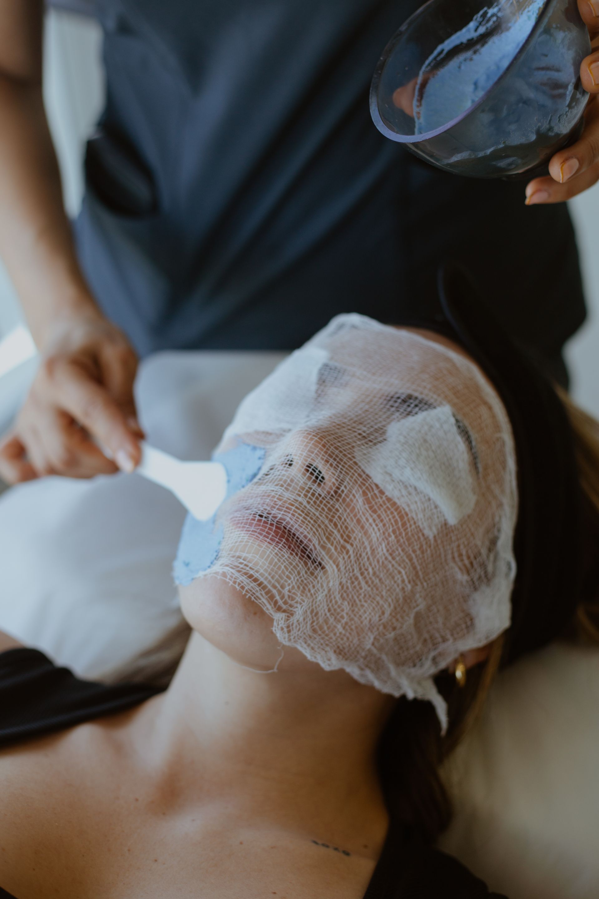 Se está aplicando una mascarilla facial de spa con una brocha a una persona tumbada.