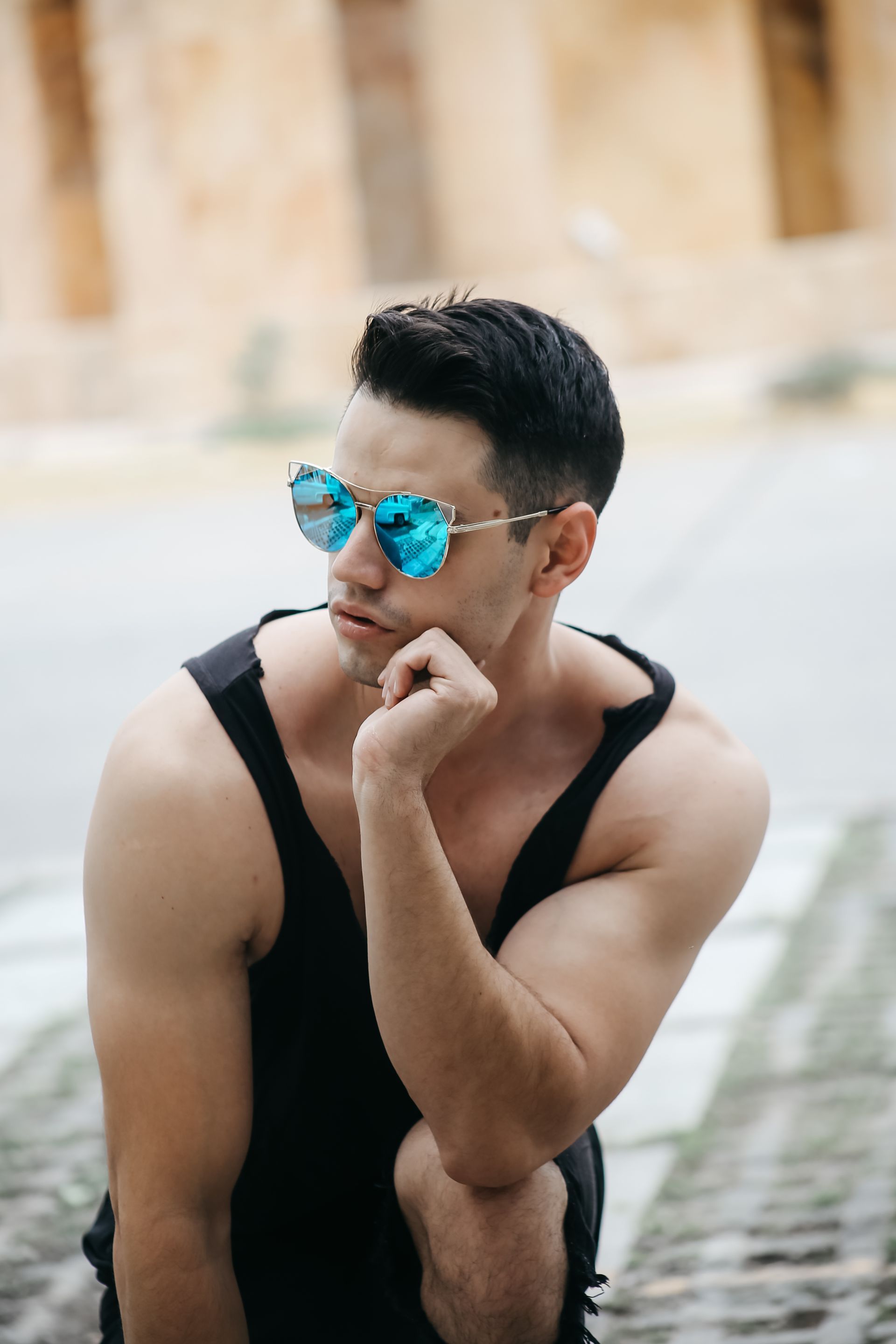 Un hombre con camiseta negra de tirantes y gafas de sol azules está agachado en un sendero de piedra, apoyando la barbilla en la mano.