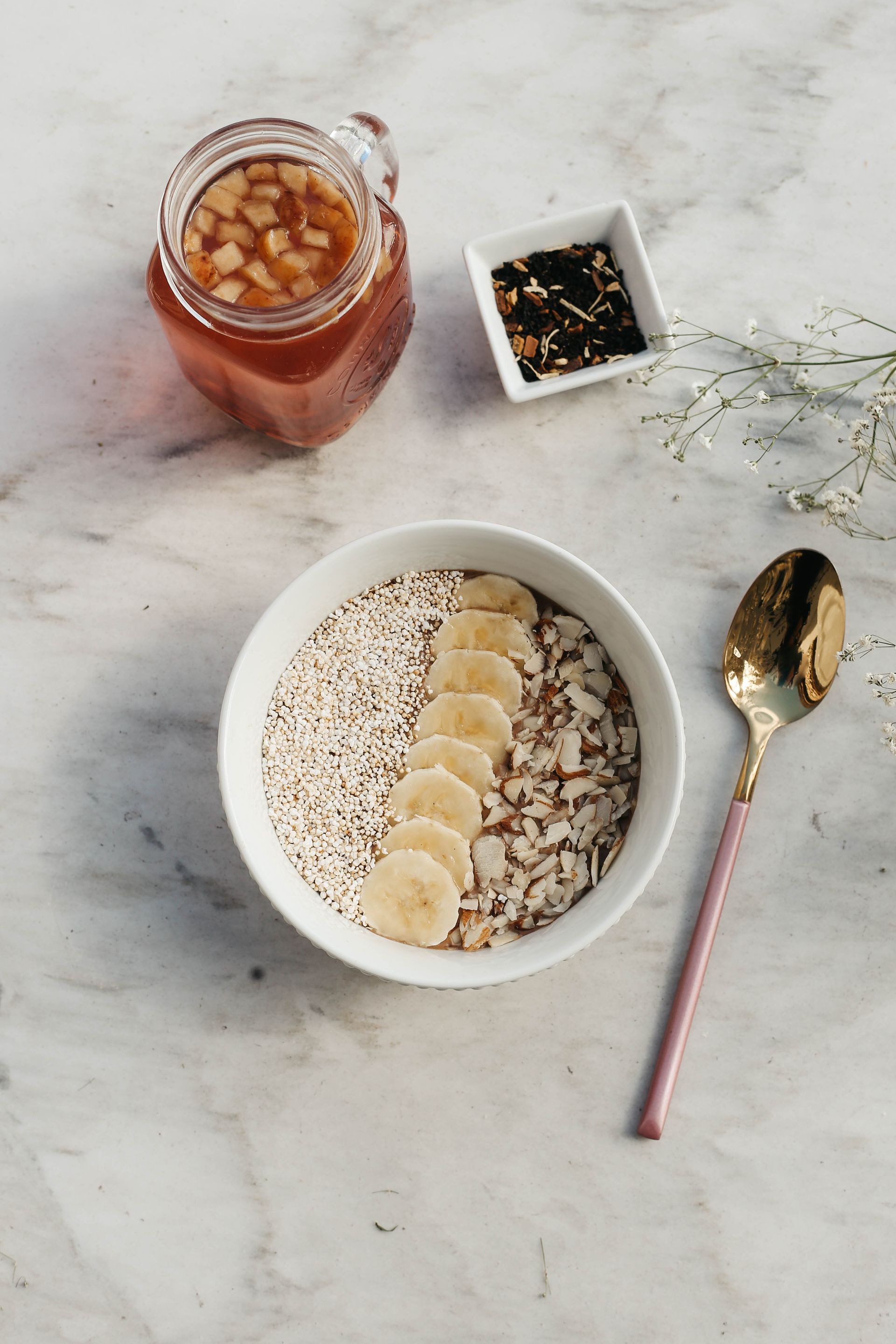 Bol de desayuno con rodajas de plátano, granola, tarro de mermelada, especiero y cuchara sobre una superficie de mármol claro.
