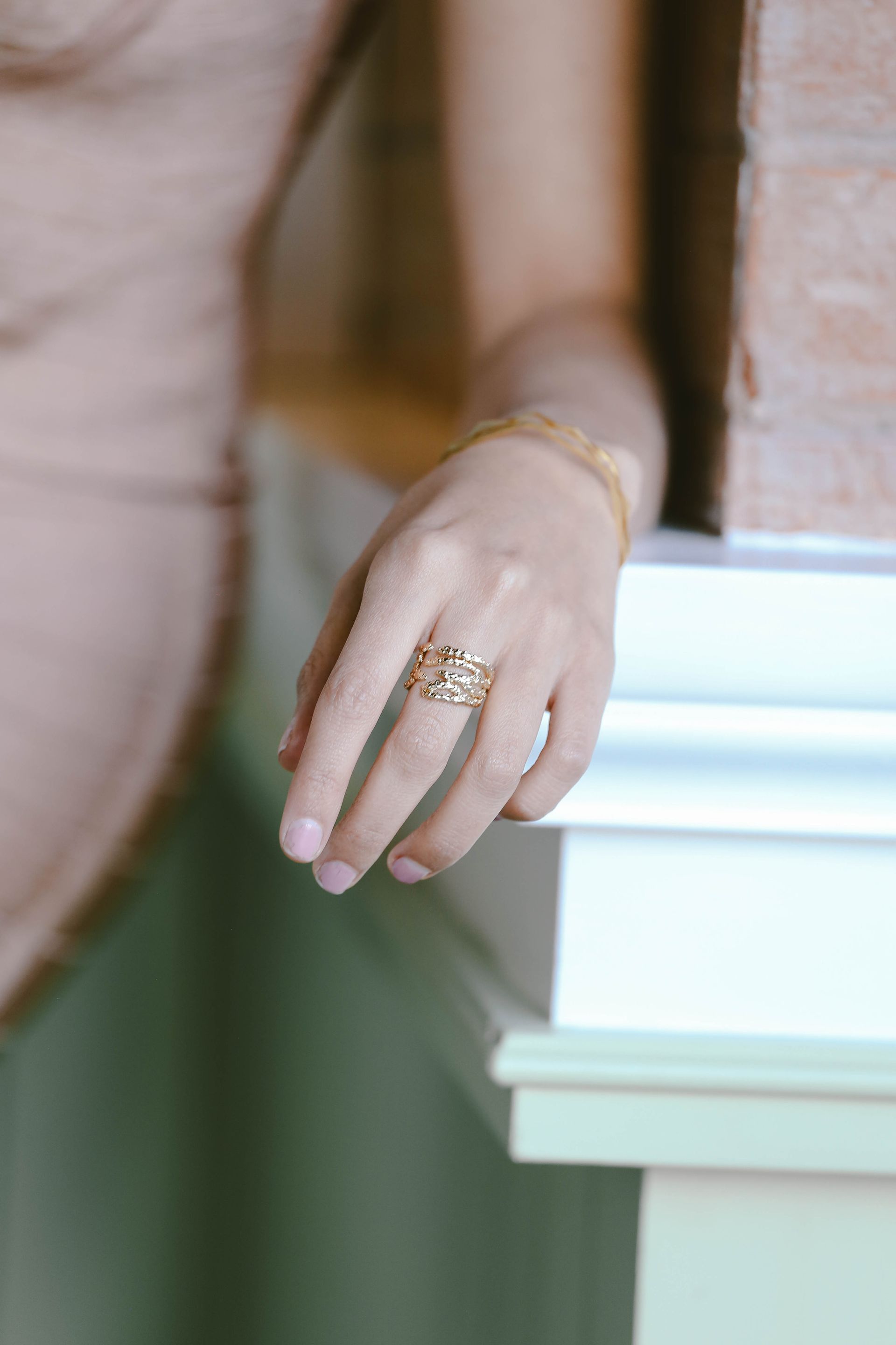 Primer plano de una mano con un anillo de oro apoyada sobre una repisa blanca, con un fondo verde suave.