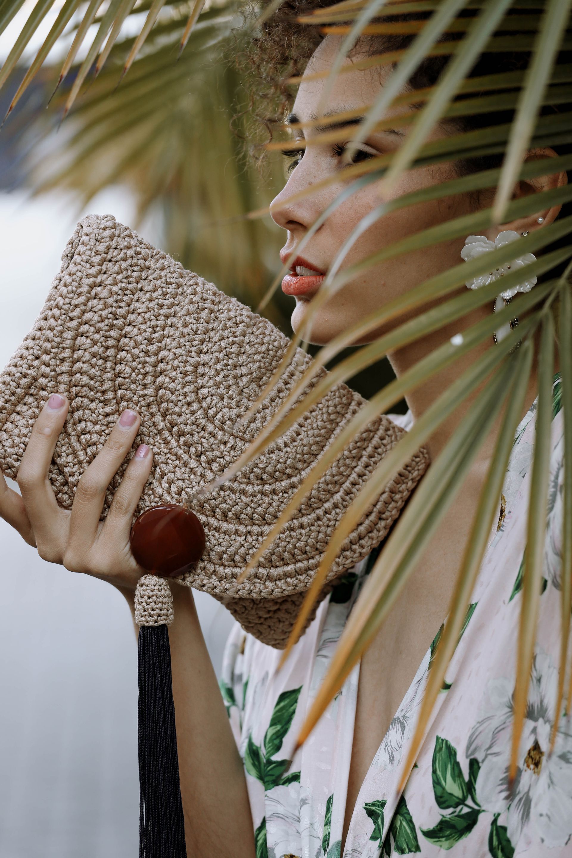Mujer con un vestido blanco estampado de flores, sosteniendo un bolso de mano tejido detrás de hojas de palmera.