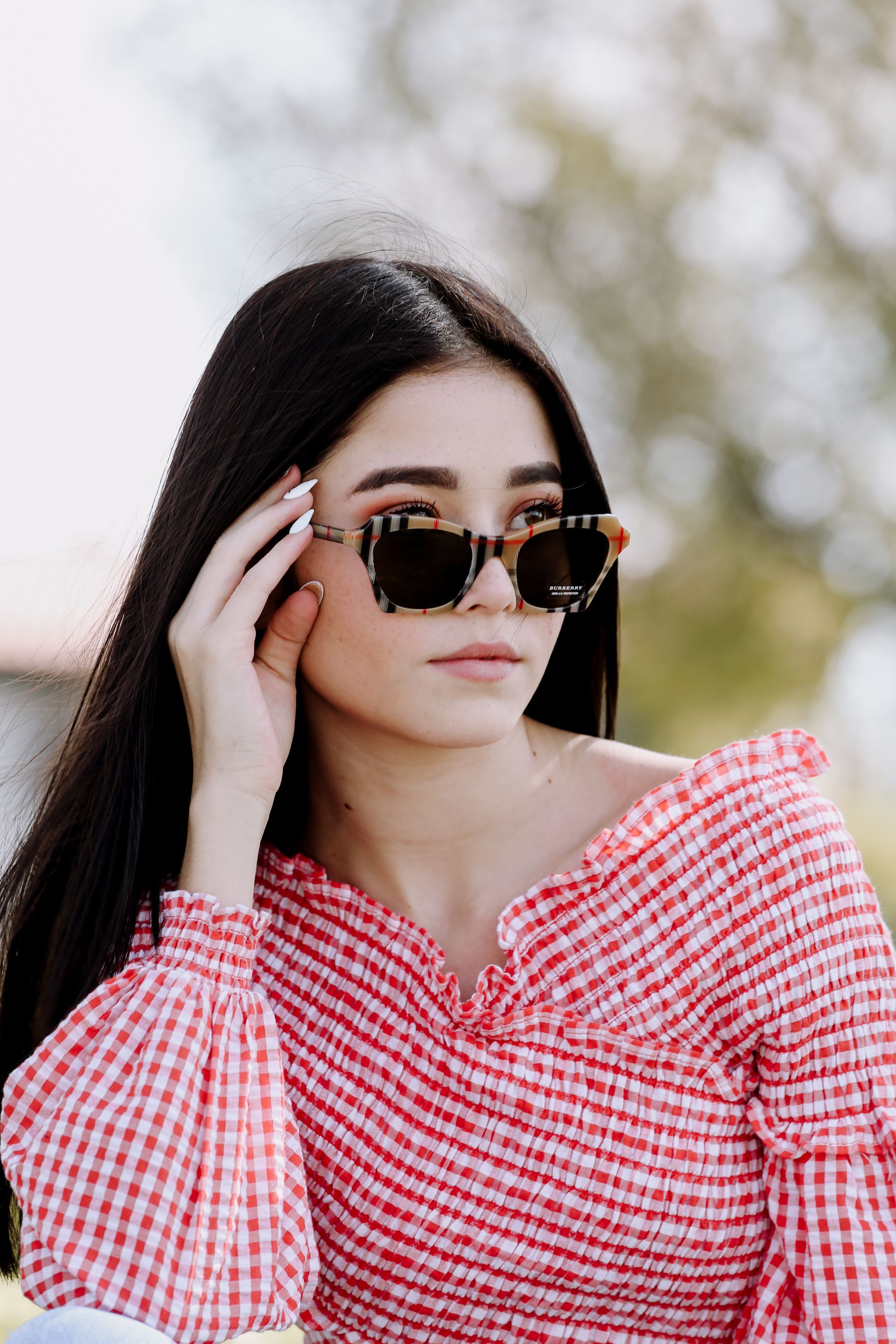 Mujer con gafas de sol arreglándose el pelo, vestida con una blusa a cuadros rojos y blancos al aire libre.