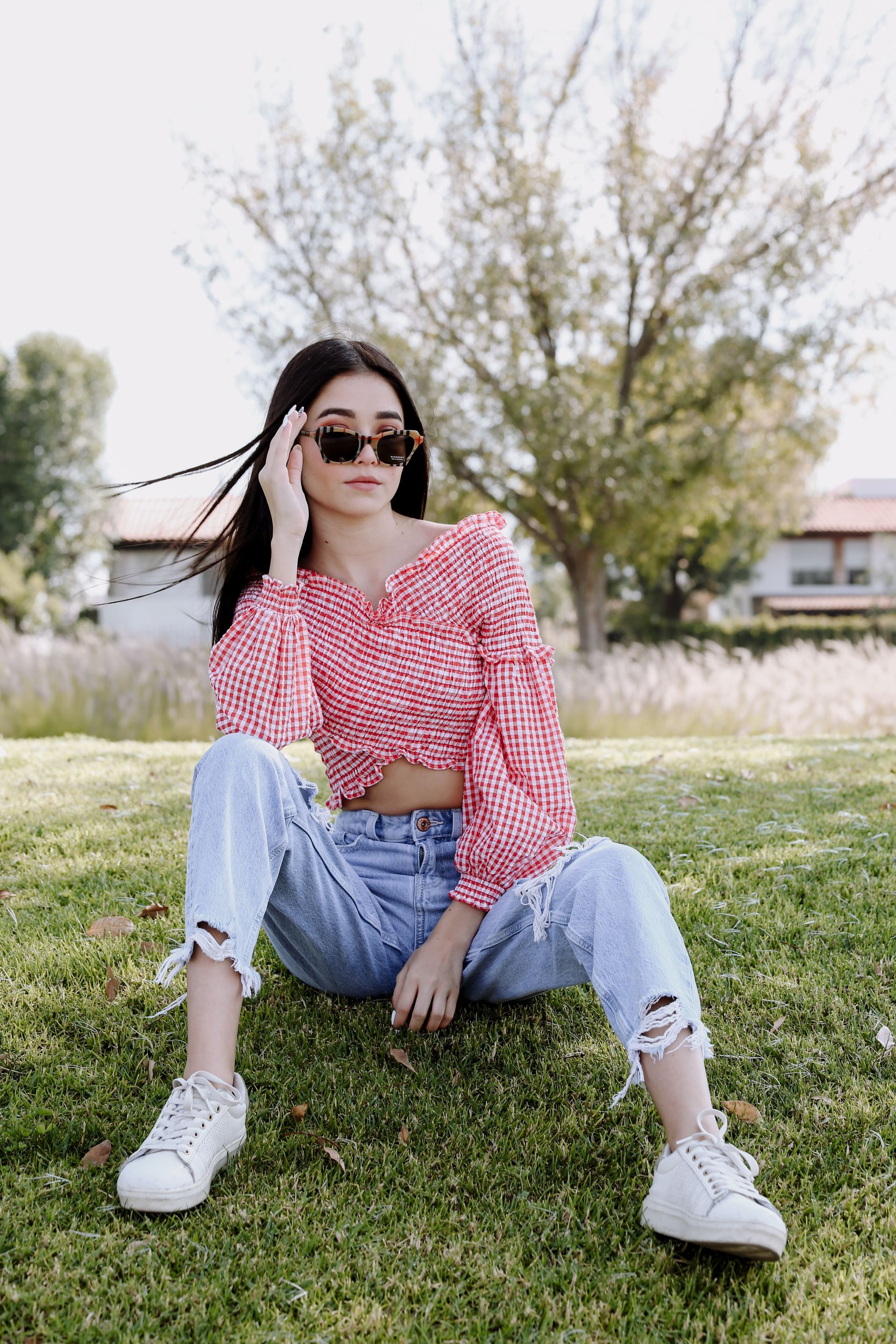 Mujer sentada en el césped con gafas de sol, blusa a cuadros roja, vaqueros claros y zapatillas blancas al aire libre.