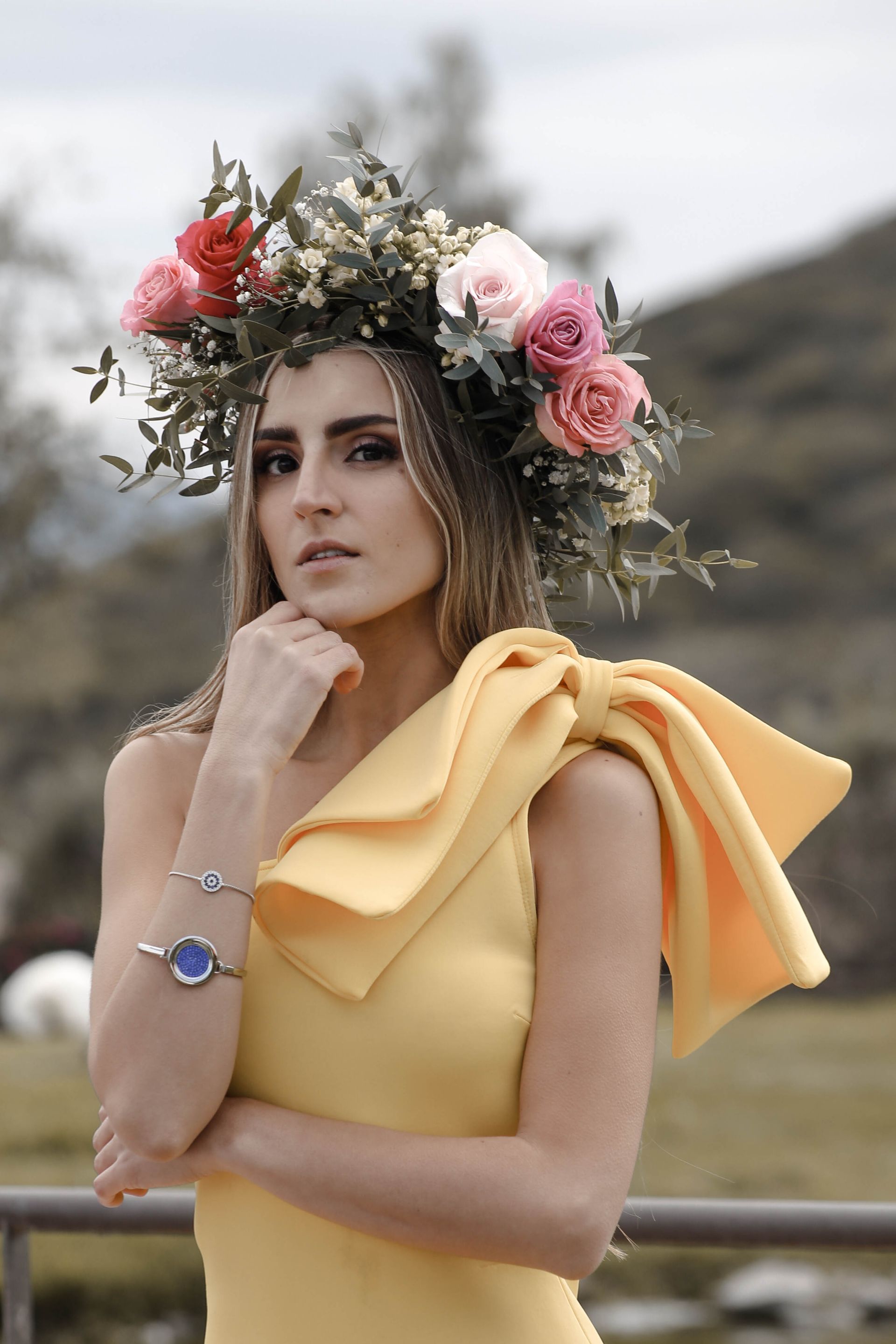Mujer con un vestido amarillo y una corona de flores, posando al aire libre con los brazos cruzados.