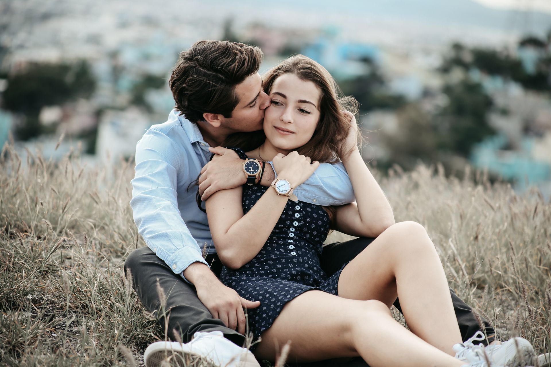 Pareja sentada en la hierba alta, abrazada y sonriendo, con un fondo exterior borroso.