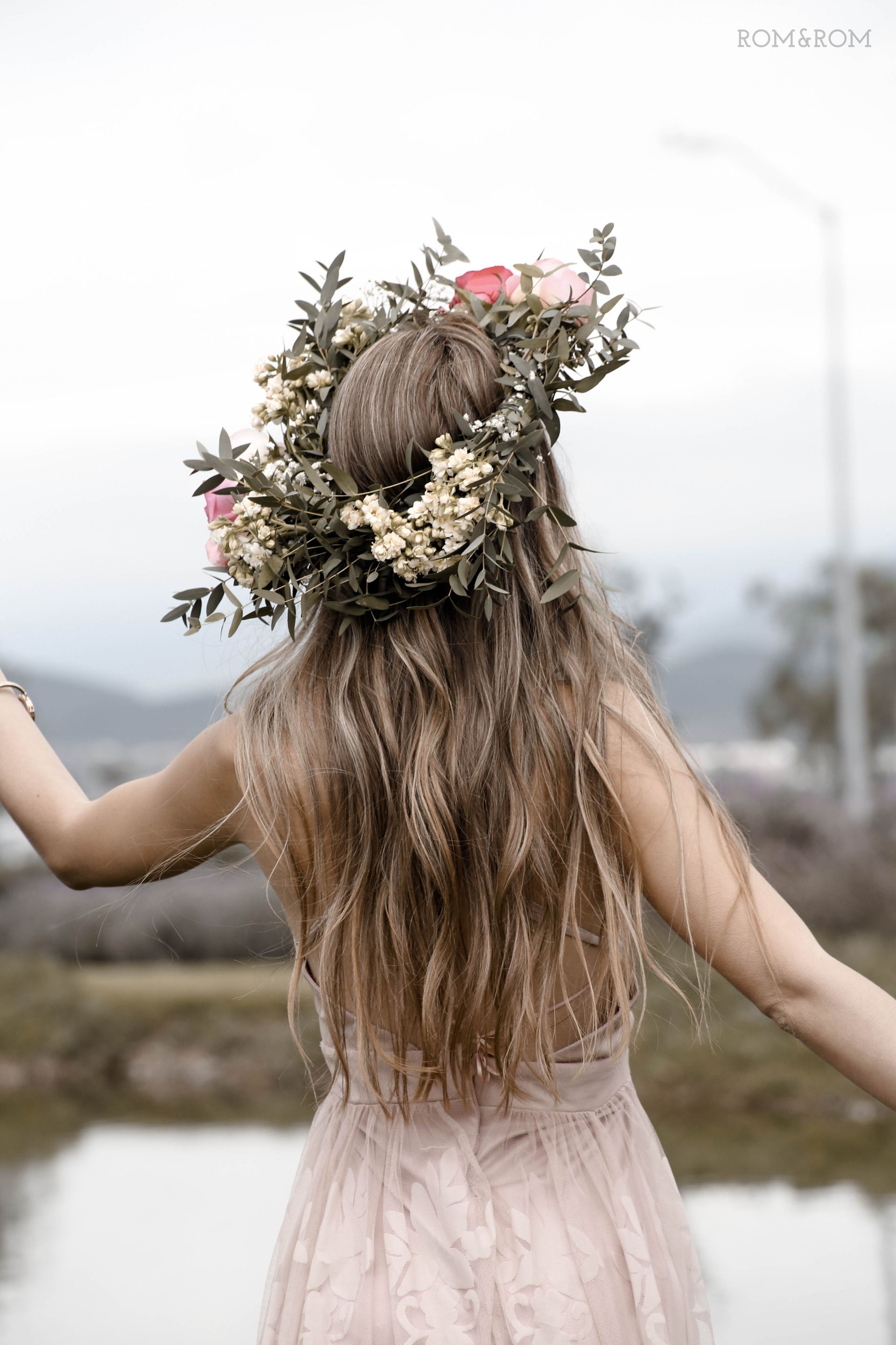 Niña con cabello largo y ondulado y corona de flores, vestida con un vestido rosa, de pie al aire libre junto a un lago.