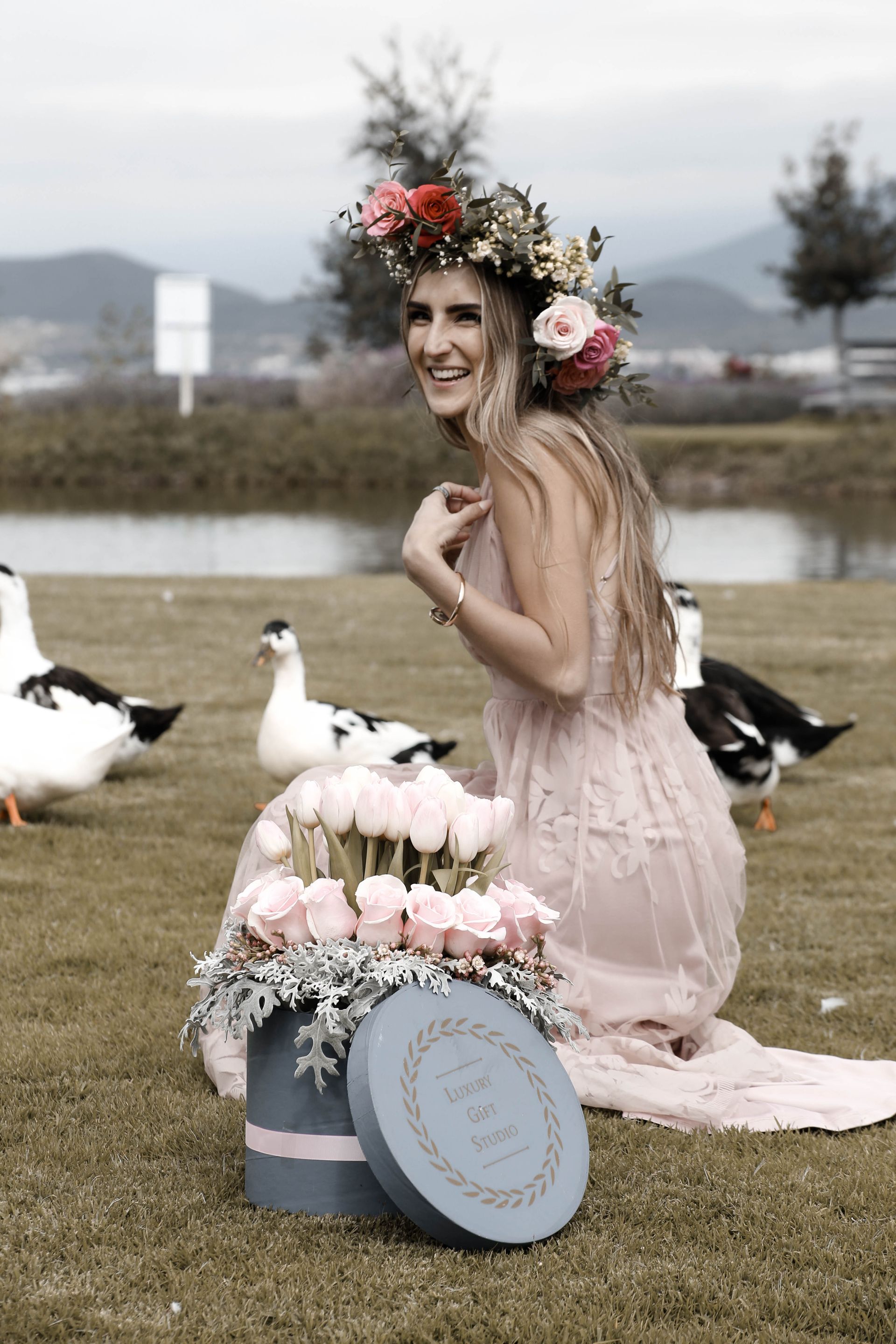 Una mujer con una corona de flores se arrodilla junto a una cesta de pastel decorada sobre el césped, rodeada de gaviotas cerca de un lago.