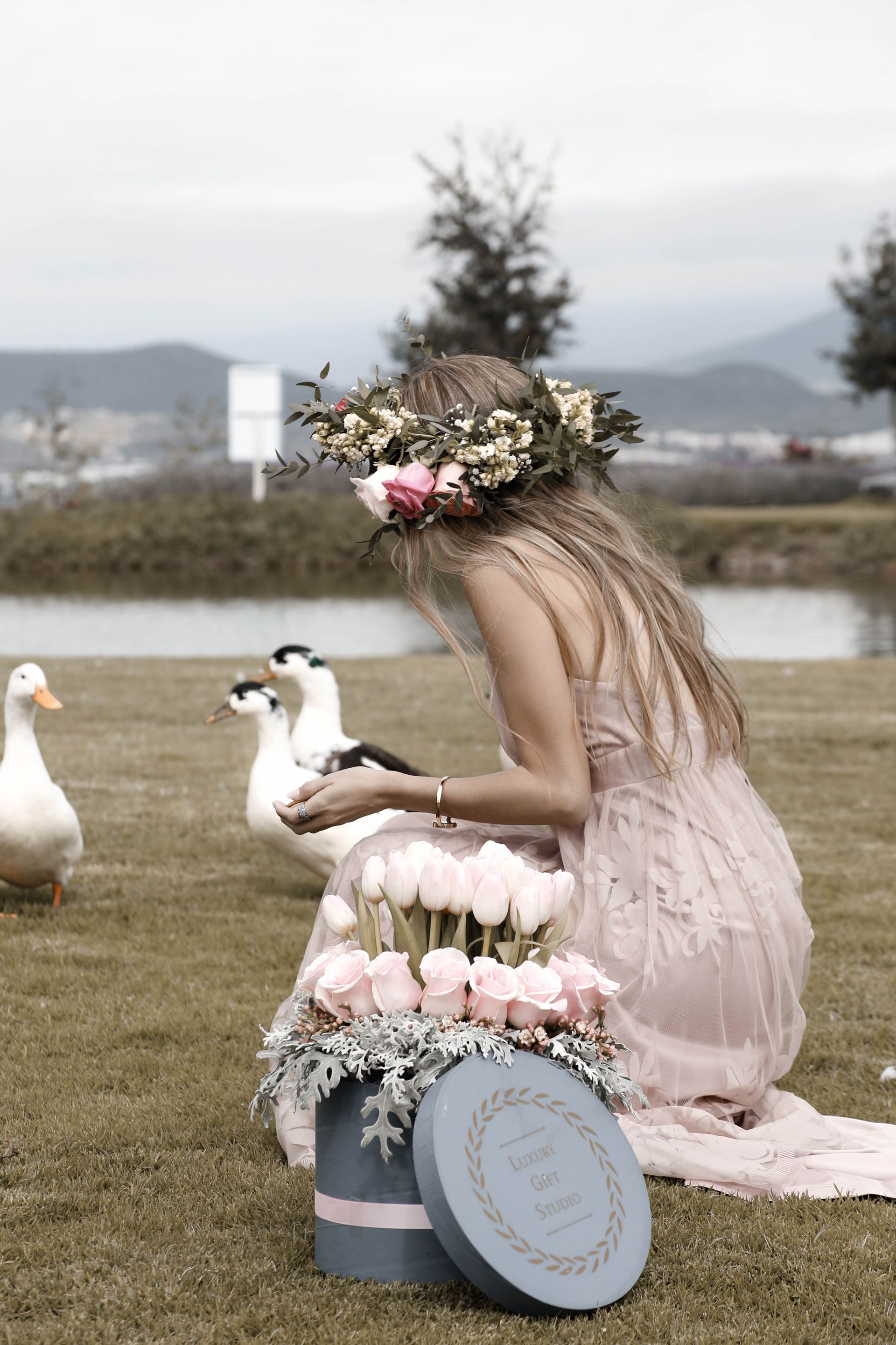 Una mujer con una corona de flores se agacha junto a unos patos al lado de una jardinera de flores de color pastel cerca de un lago.