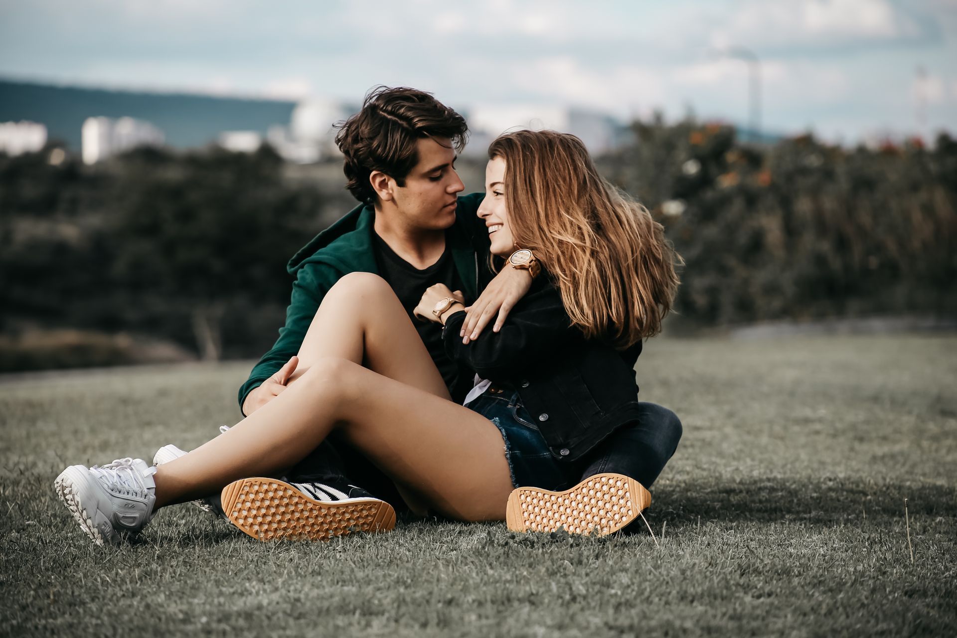 Una pareja sentada en el césped al aire libre, inclinada una hacia la otra y sonriéndose.