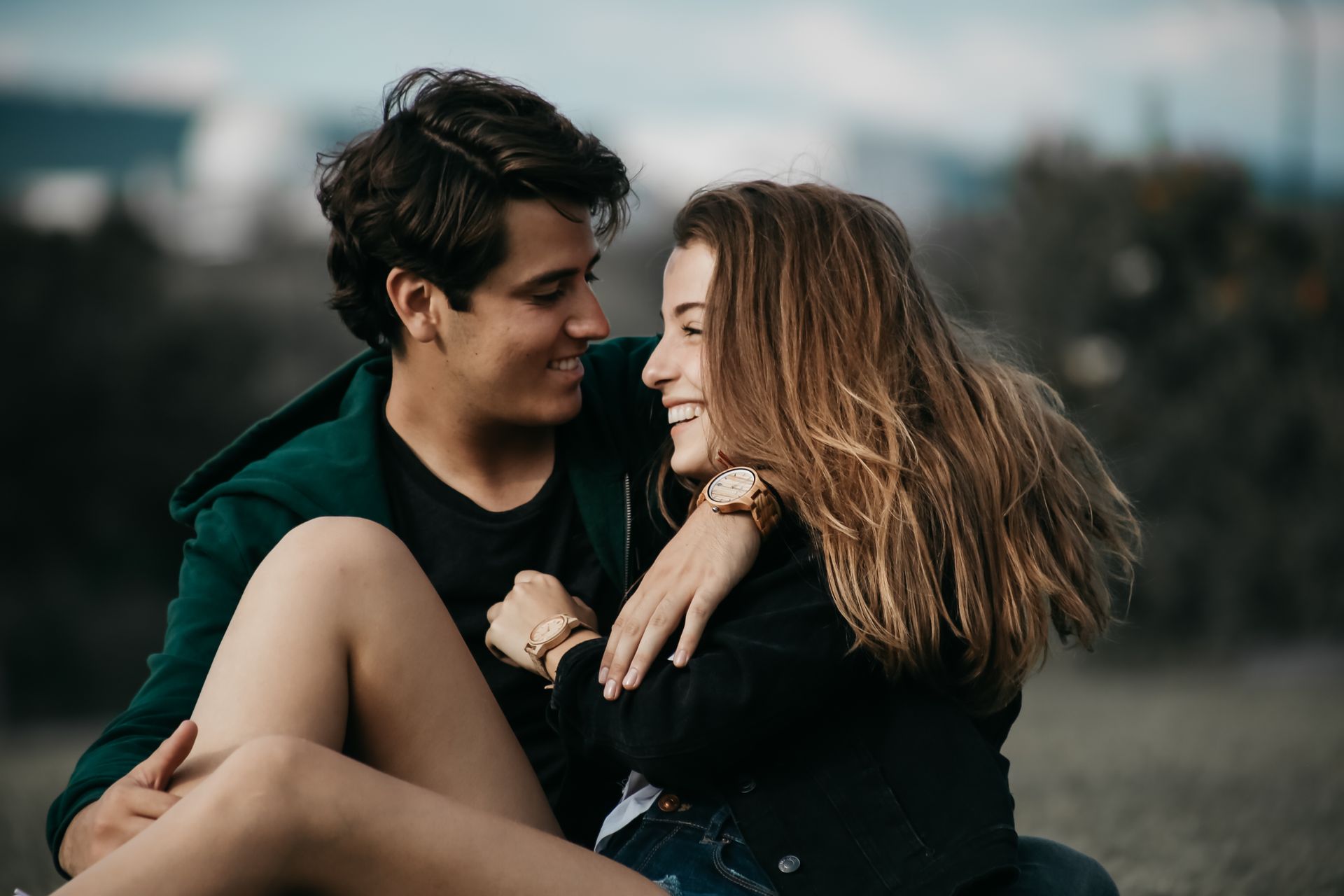 Una pareja sentada muy cerca el uno del otro al aire libre, sonriendo y mirándose bajo una luz tenue.