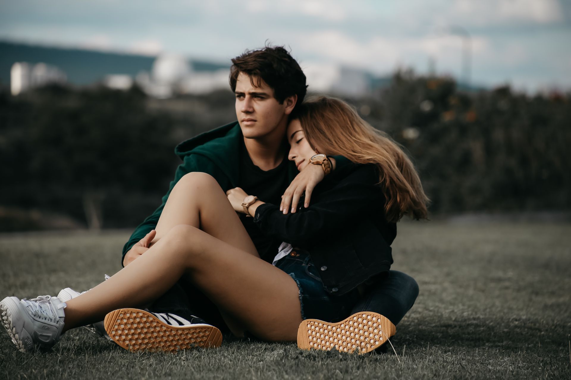 Dos personas sentadas en el césped al aire libre, una apoyada en la otra en un entorno nublado similar a un parque.