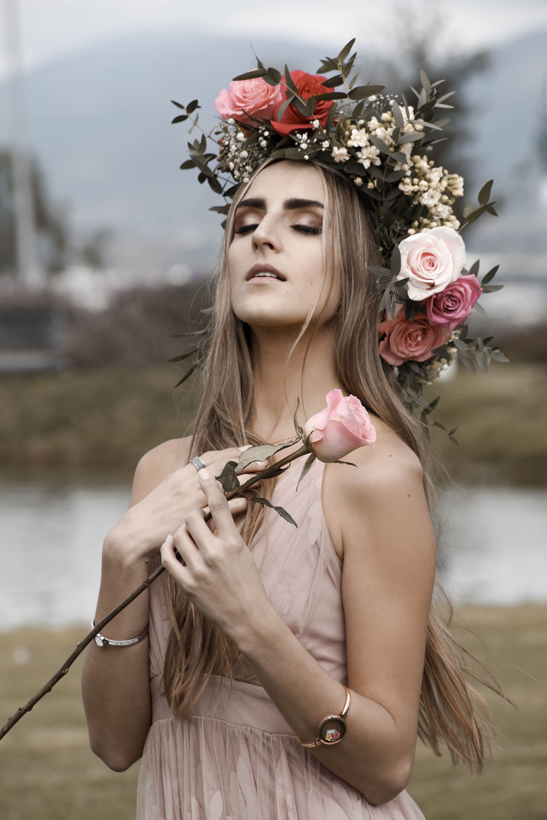 Mujer con corona de flores y vestido rosa, sosteniendo una rosa rosa en un entorno brumoso al aire libre.