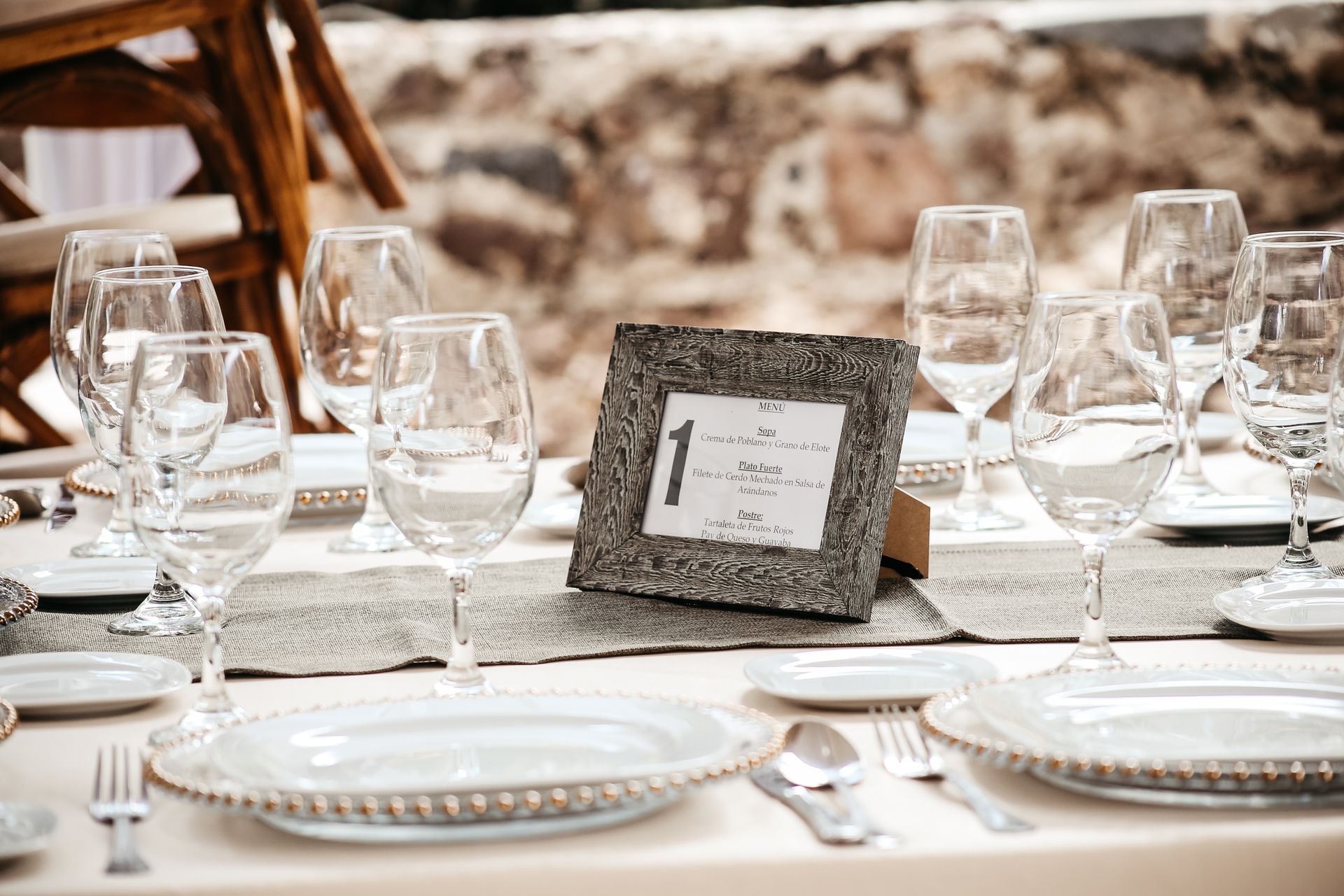 Elegante mesa de exterior con platos blancos, cristalería y un centro de mesa con el número de mesa enmarcado.