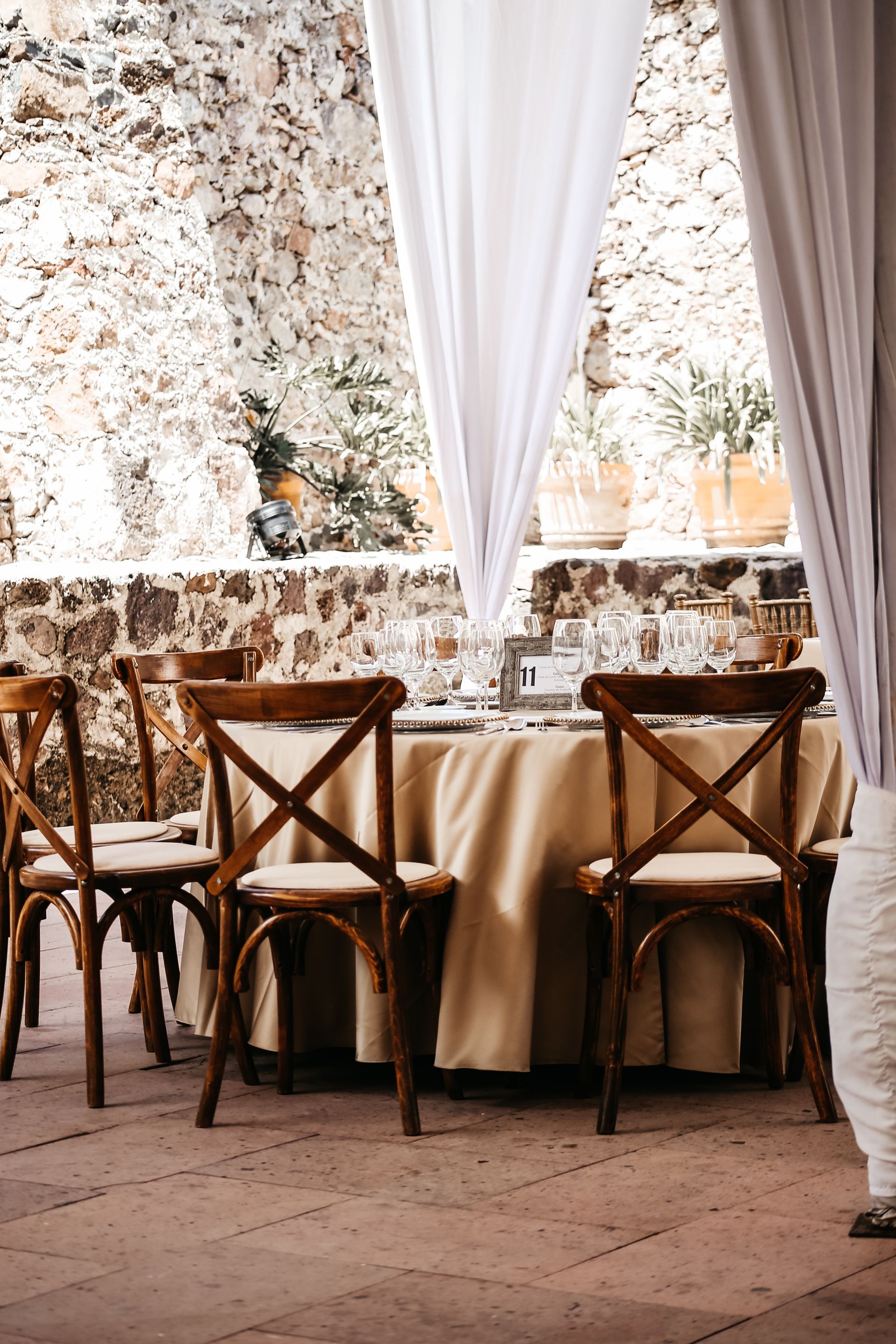 Elegante comedor al aire libre con mesas redondas, sillas de madera y cortinas blancas.