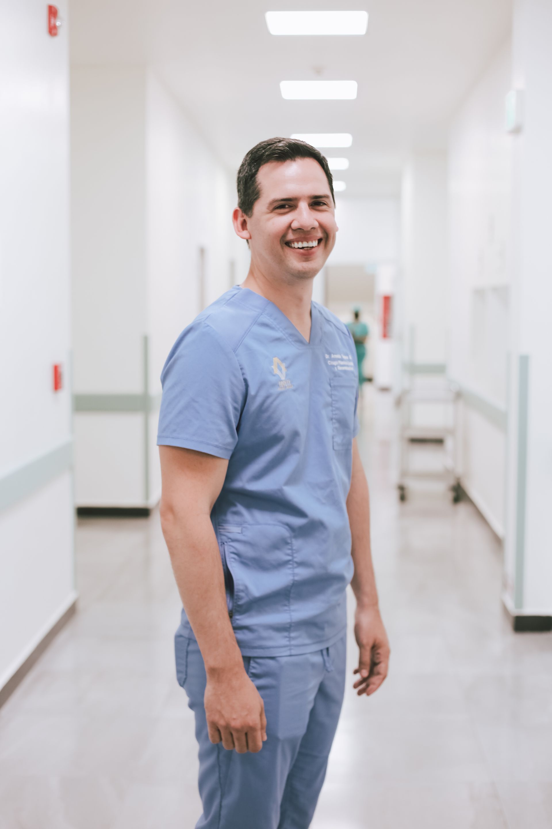 Hombre con uniforme médico azul claro sonriendo en un pasillo luminoso del hospital.