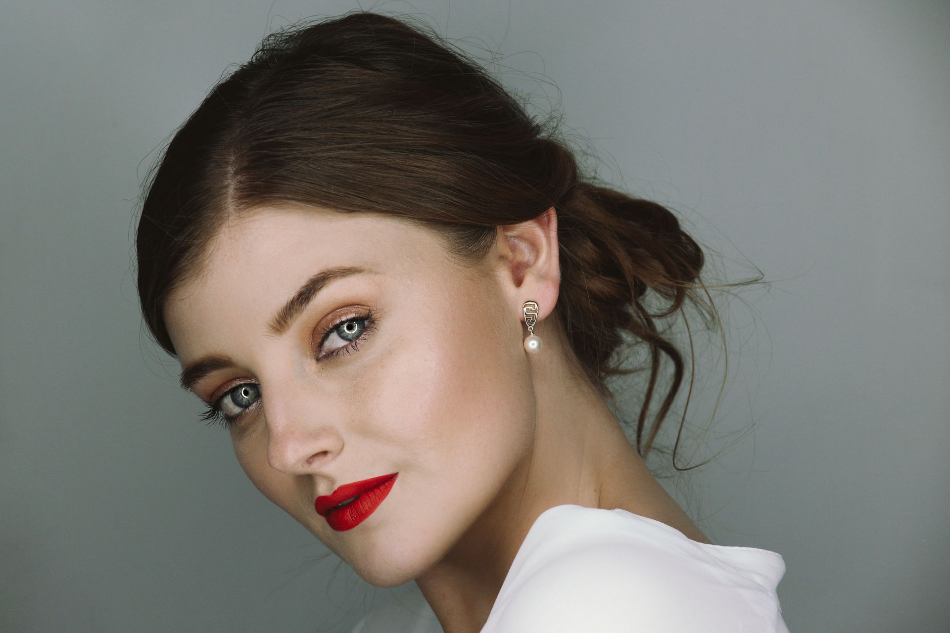 Retrato de una mujer de cabello oscuro, lápiz labial rojo y pendientes, de perfil sobre un fondo gris.