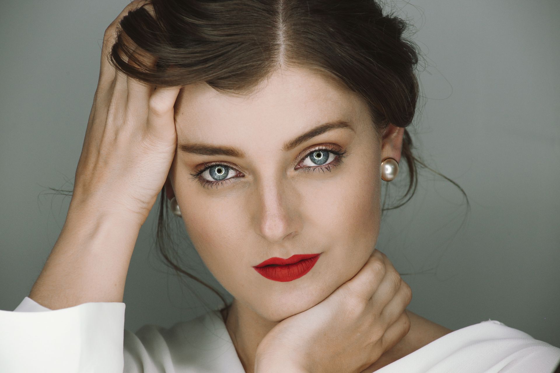 Retrato de una mujer con cabello oscuro, ojos azules, lápiz labial rojo y pendientes de perlas sobre un fondo gris.