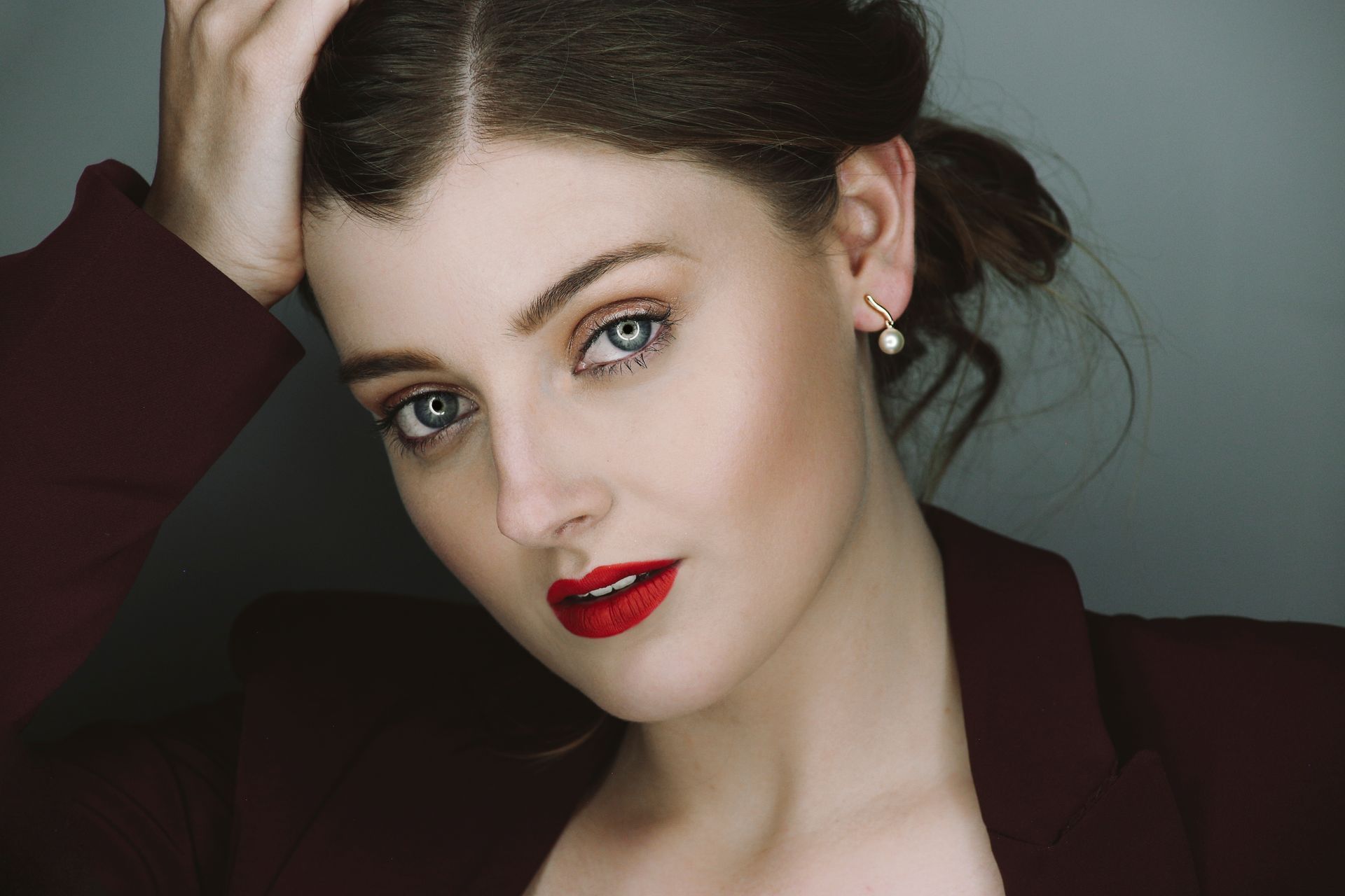 Retrato en primer plano de una mujer con cabello oscuro, lápiz labial rojo y una chaqueta granate sobre un fondo gris suave.