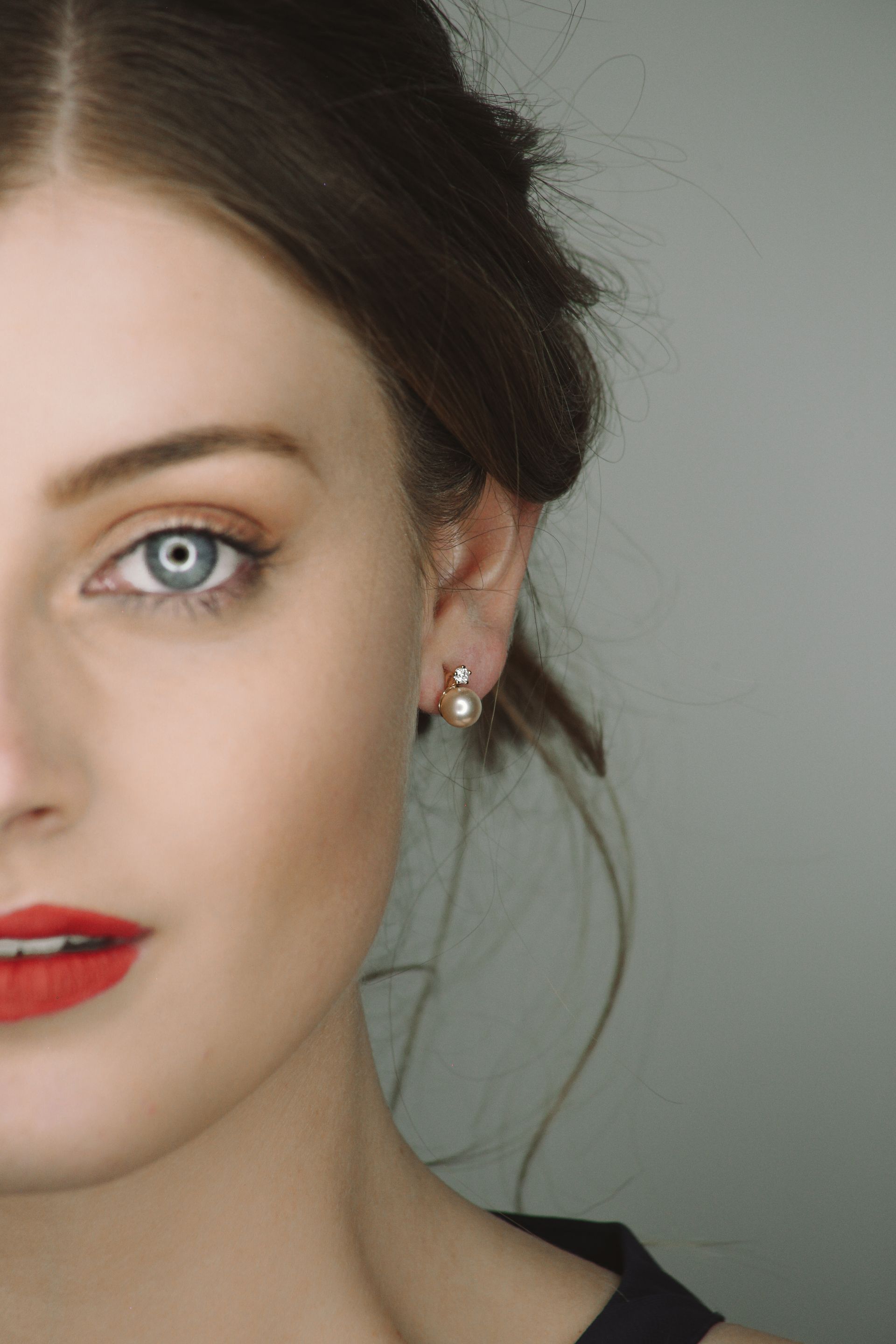 Primer plano del rostro de una mujer con lápiz labial rojo y pendientes de perlas sobre un fondo gris.