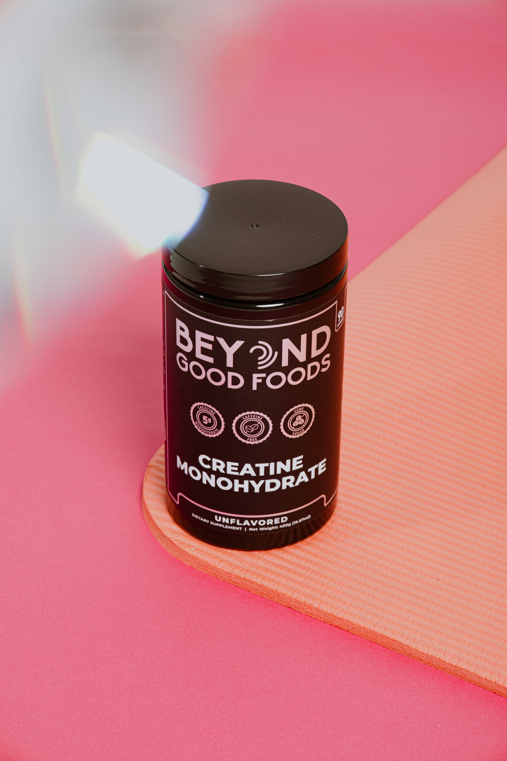 Frasco negro de monohidrato de creatina de Beyond Good Foods sobre una superficie rosa con un fondo de tela color melocotón.