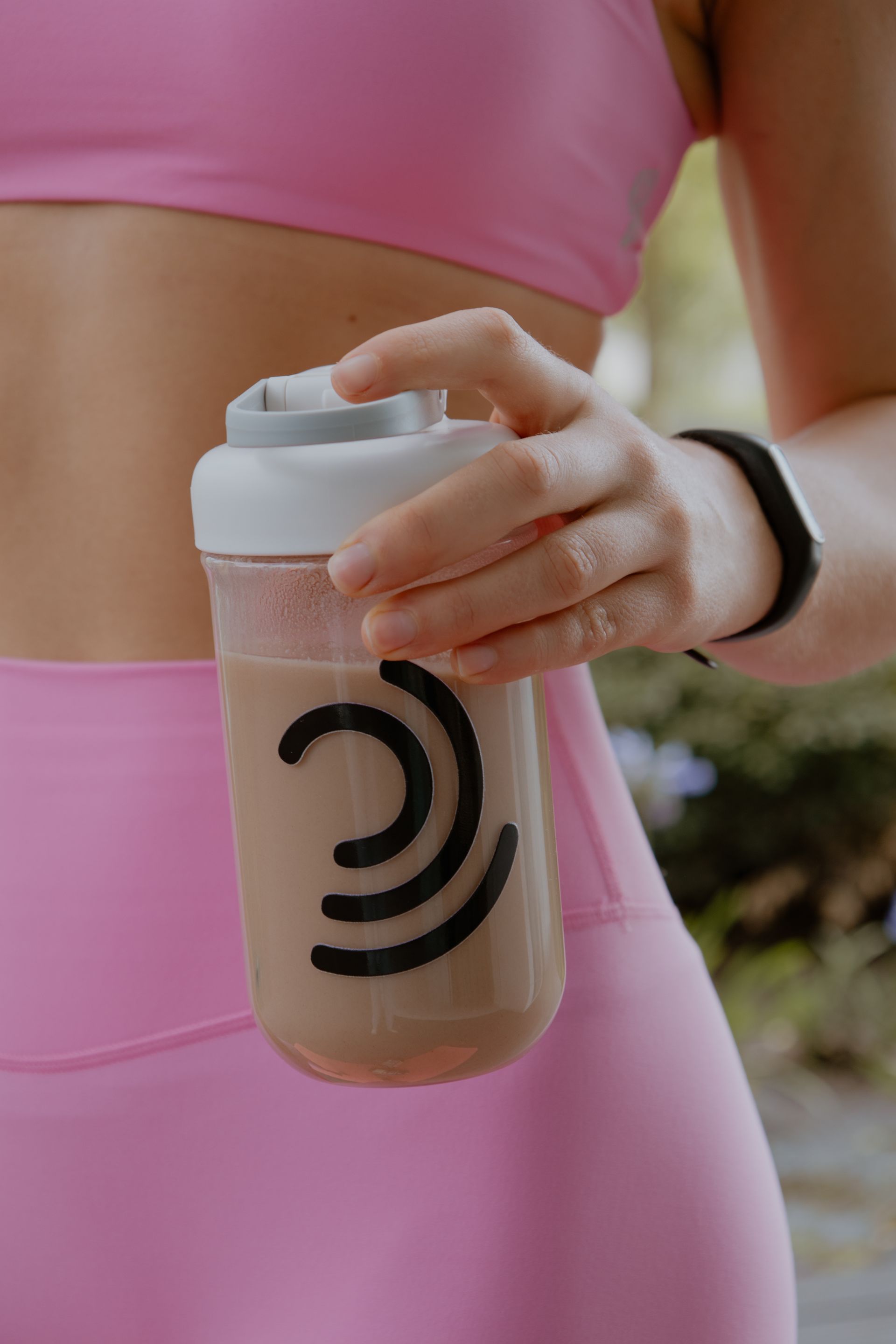 Persona con ropa deportiva rosa sosteniendo una botella mezcladora color canela con un logotipo negro al aire libre