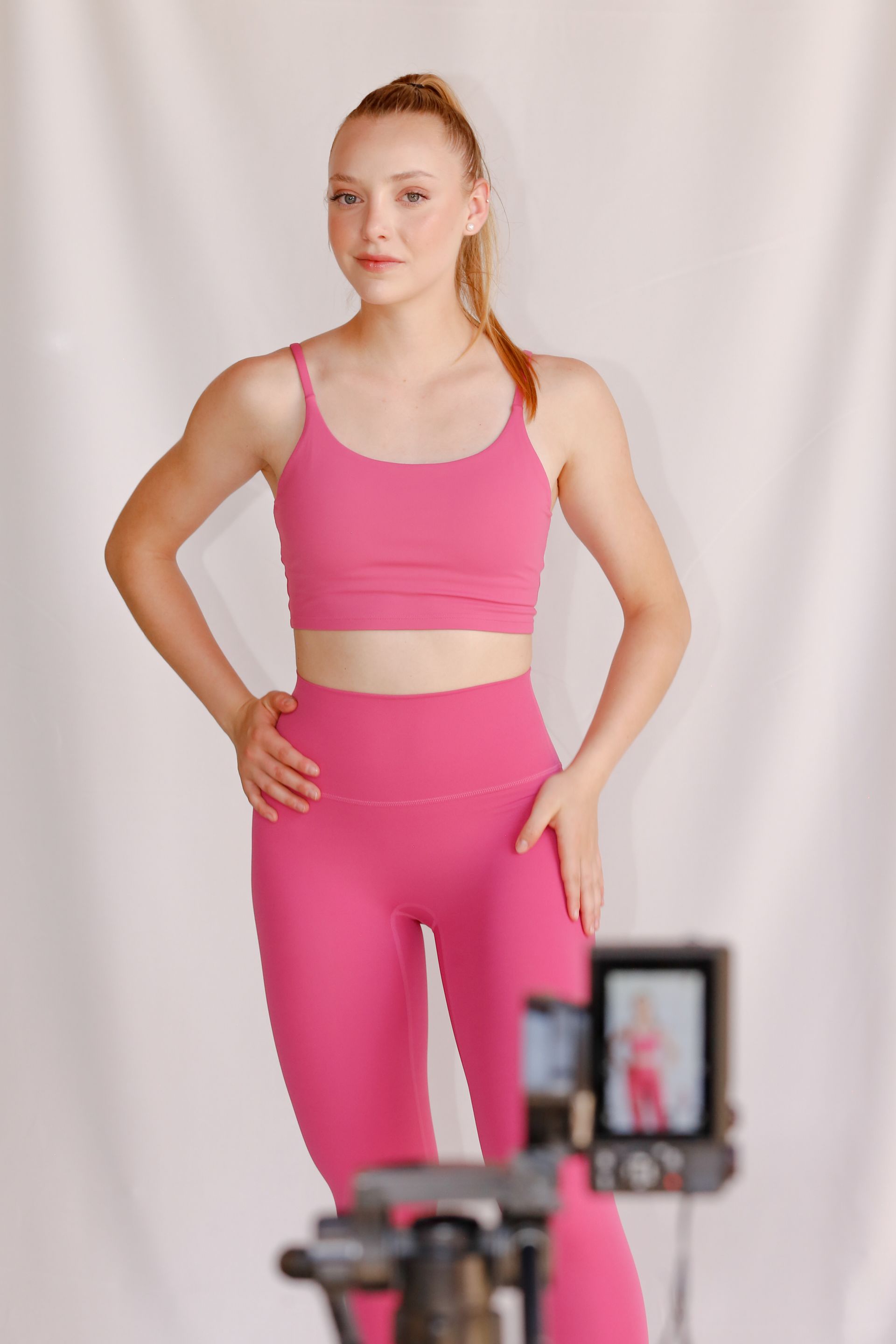 Mujer con ropa deportiva rosa posando con las manos en las caderas, fotografiada con un fondo claro.
