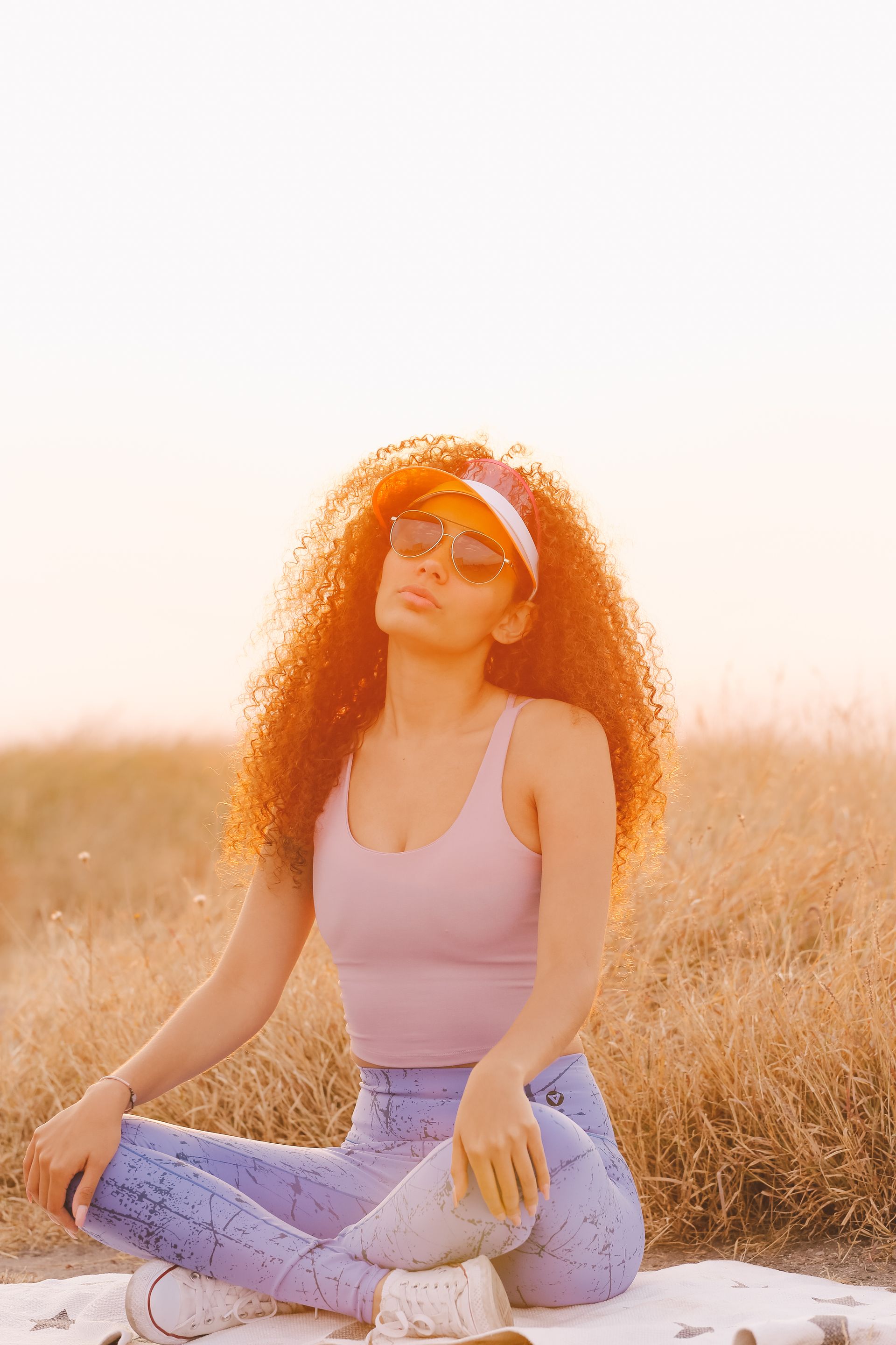 Mujer sentada con las piernas cruzadas en un campo soleado, vestida con una blusa rosa, vaqueros y gafas de sol.