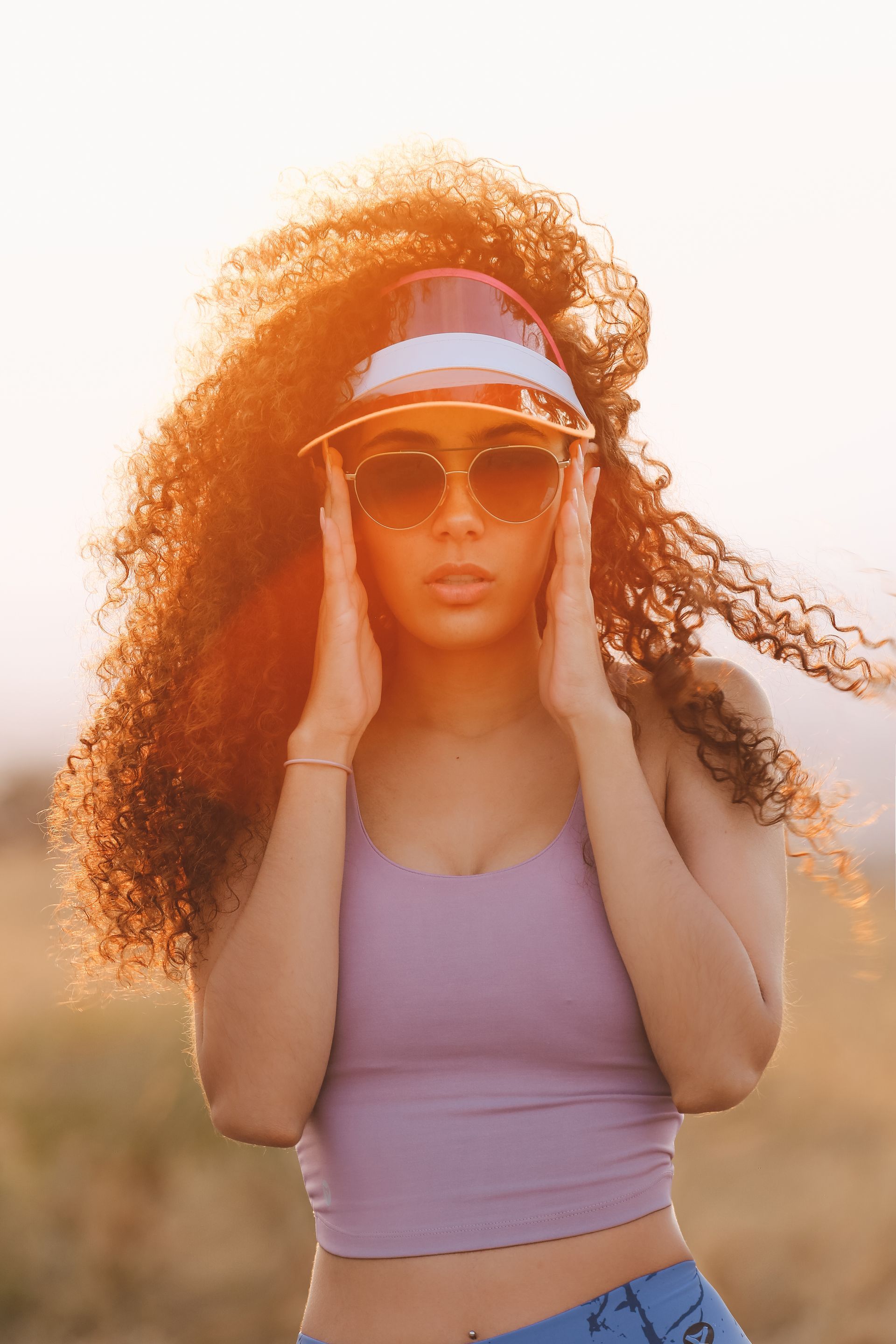 Mujer con blusa morada y gafas de sol, sosteniendo un sombrero al aire libre bajo la cálida luz del atardecer.