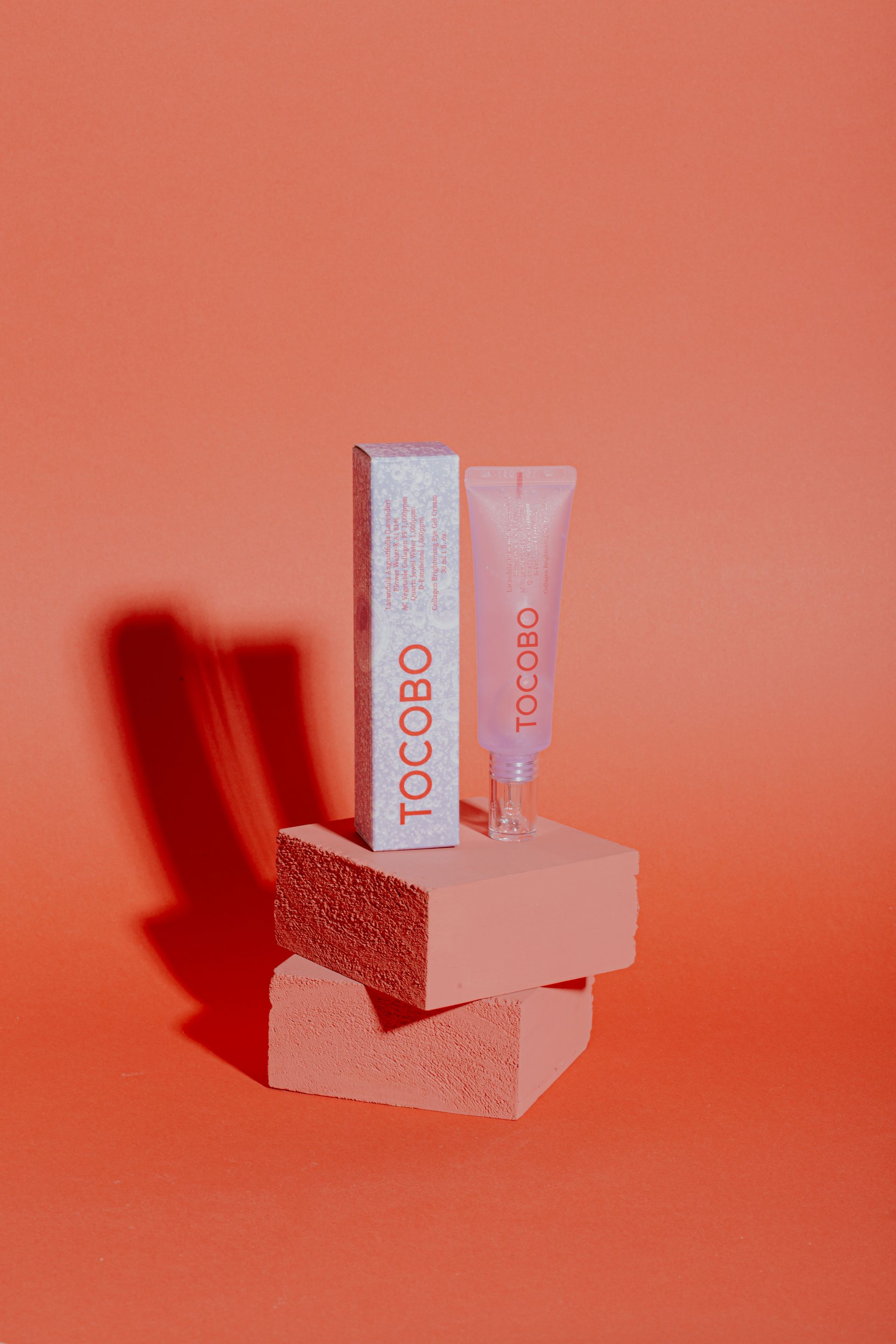 Dos tubos de cosméticos sobre un bloque texturizado, iluminados en naranja cálido con un fondo de sombra roja.