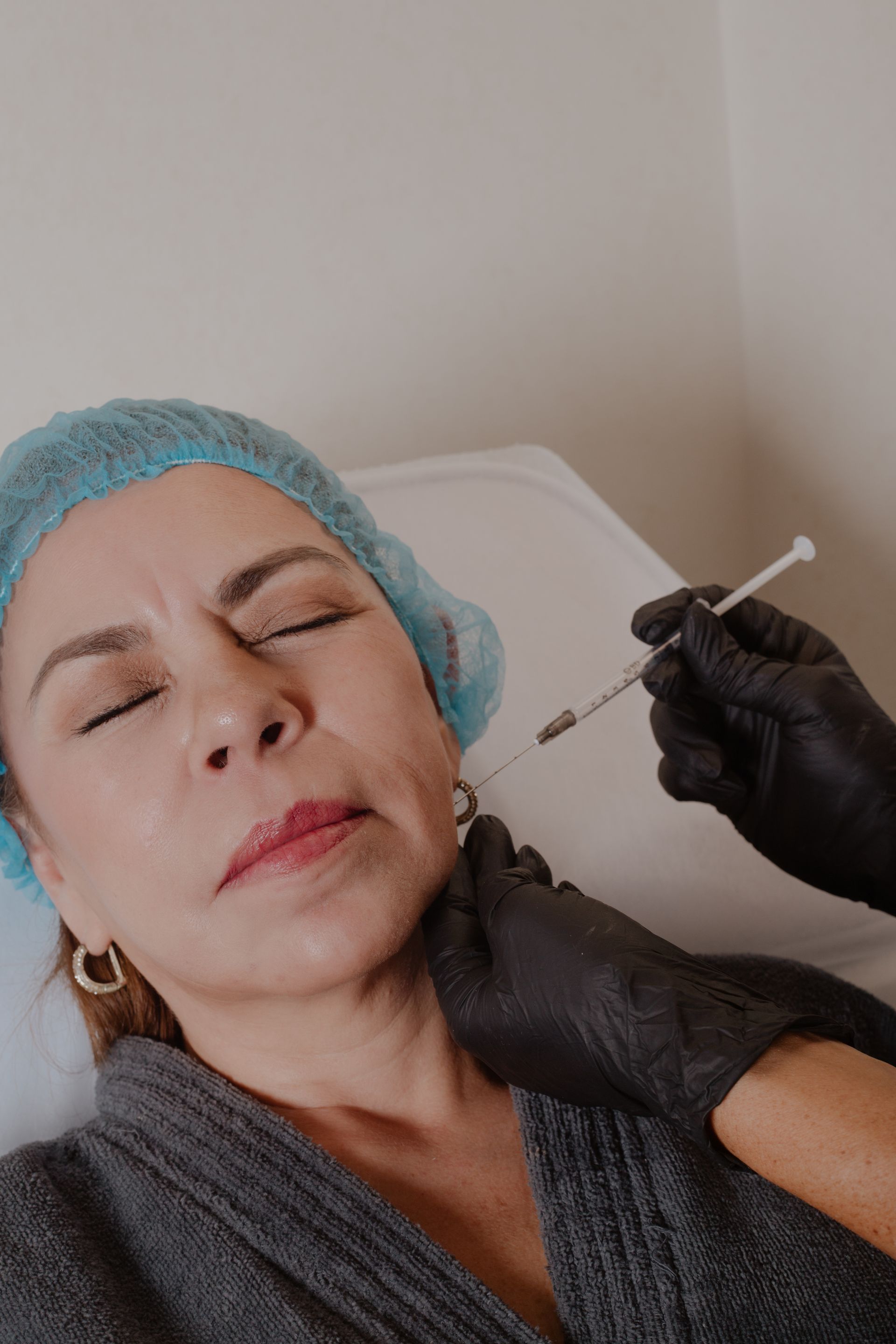 Una clienta recibe inyecciones faciales de un profesional que lleva guantes negros y una gorra azul para cubrirse el cabello.