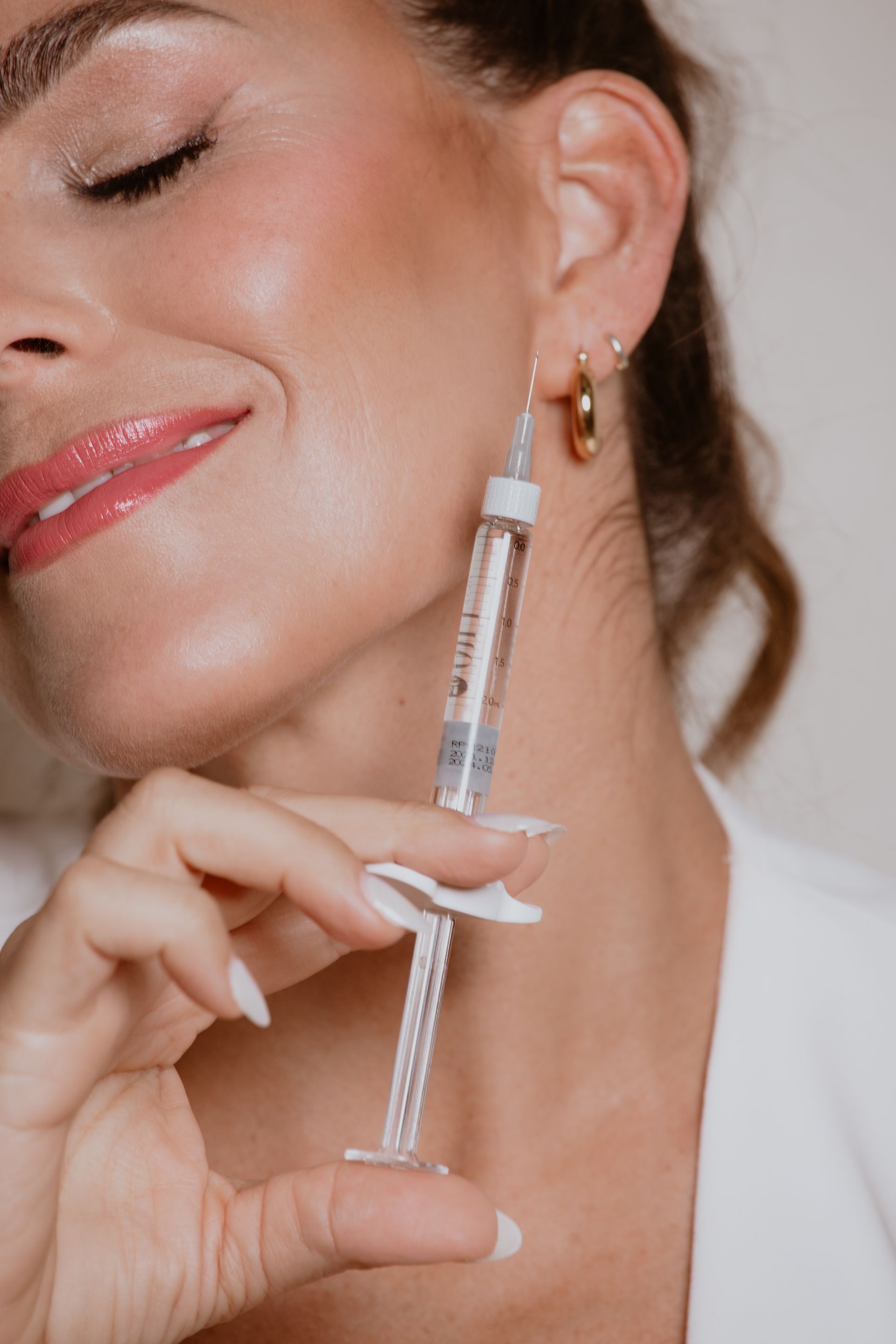 ROM&ROM  Mujer aplicándose brillo labial con un aplicador de tubo transparente, con los ojos cerrados y luciendo pendientes de aro dorados.