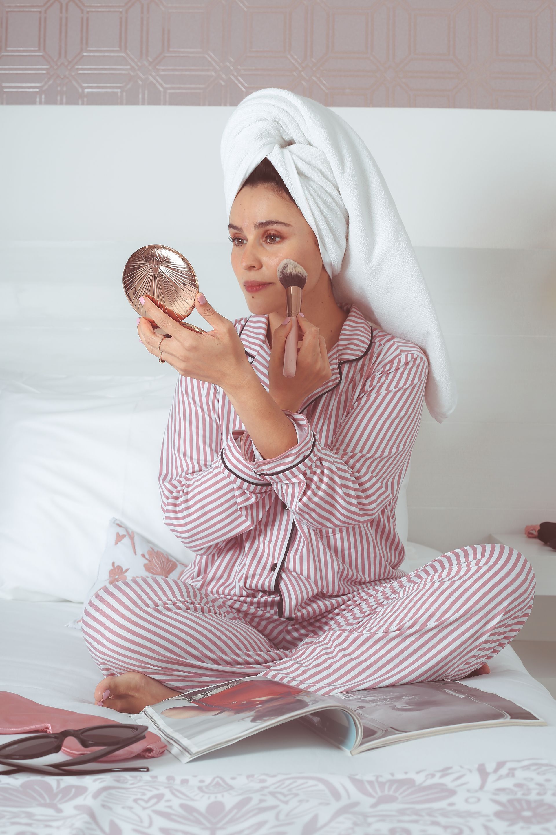 Mujer con pijama de rayas rosas aplicándose maquillaje mientras sostiene un espejo de bolsillo sobre una cama.