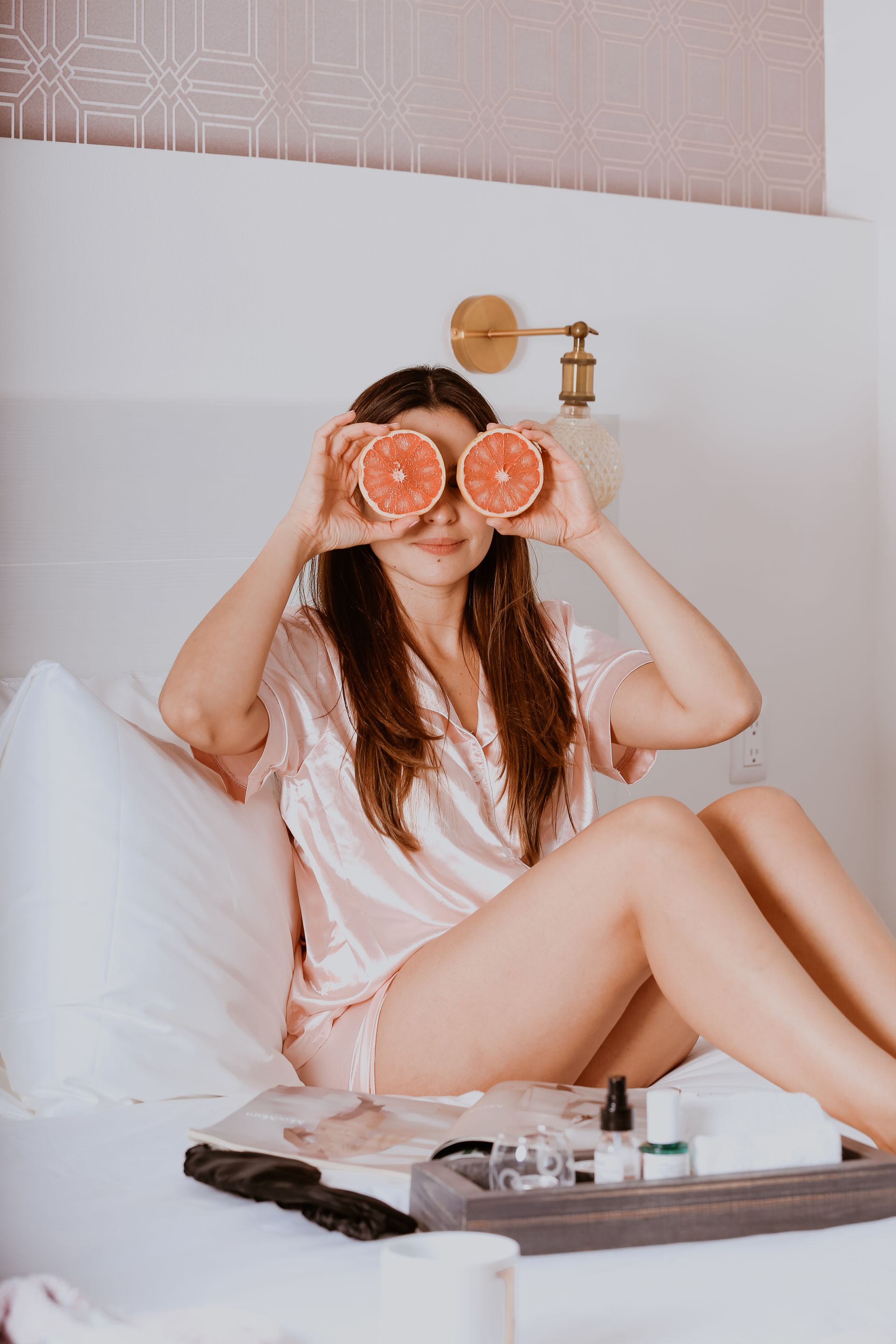 Mujer en la cama sosteniendo mitades de pomelo sobre sus ojos en una luminosa escena de spa en un dormitorio.