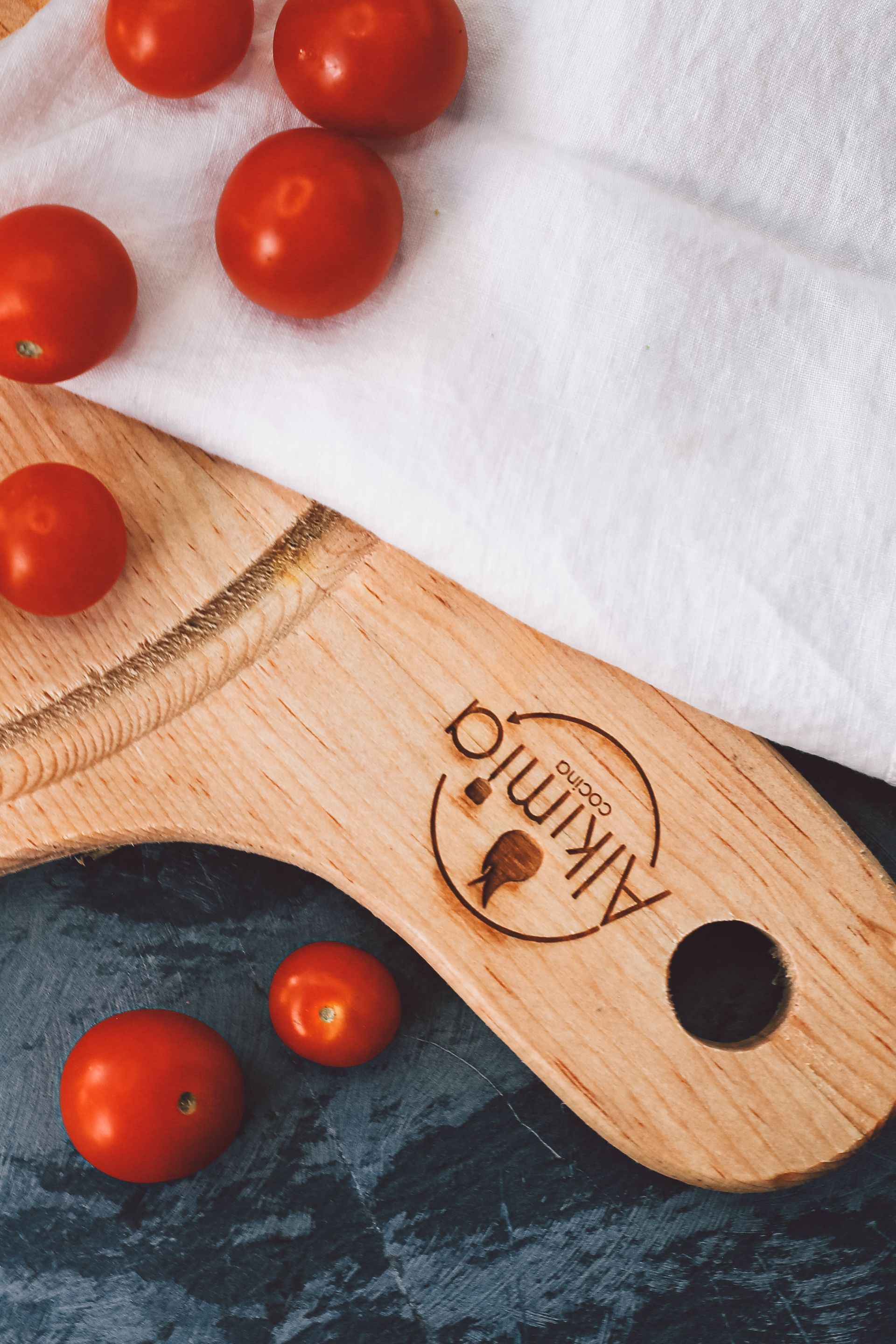 Tomates cherry sobre una tabla de cortar de madera junto a un paño blanco sobre una superficie oscura.