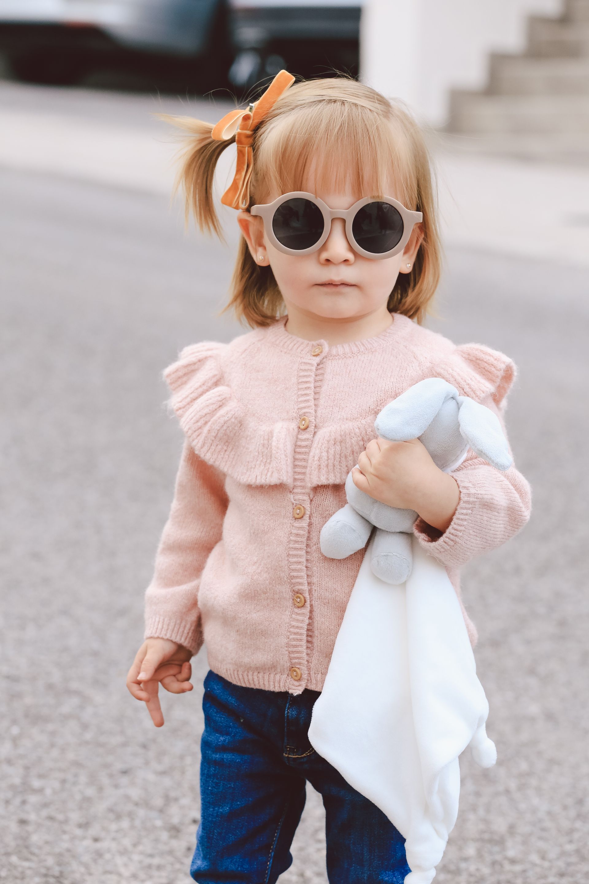 Un niño pequeño con un suéter rosa y gafas de sol sostiene un peluche, de pie al aire libre en una calle.
