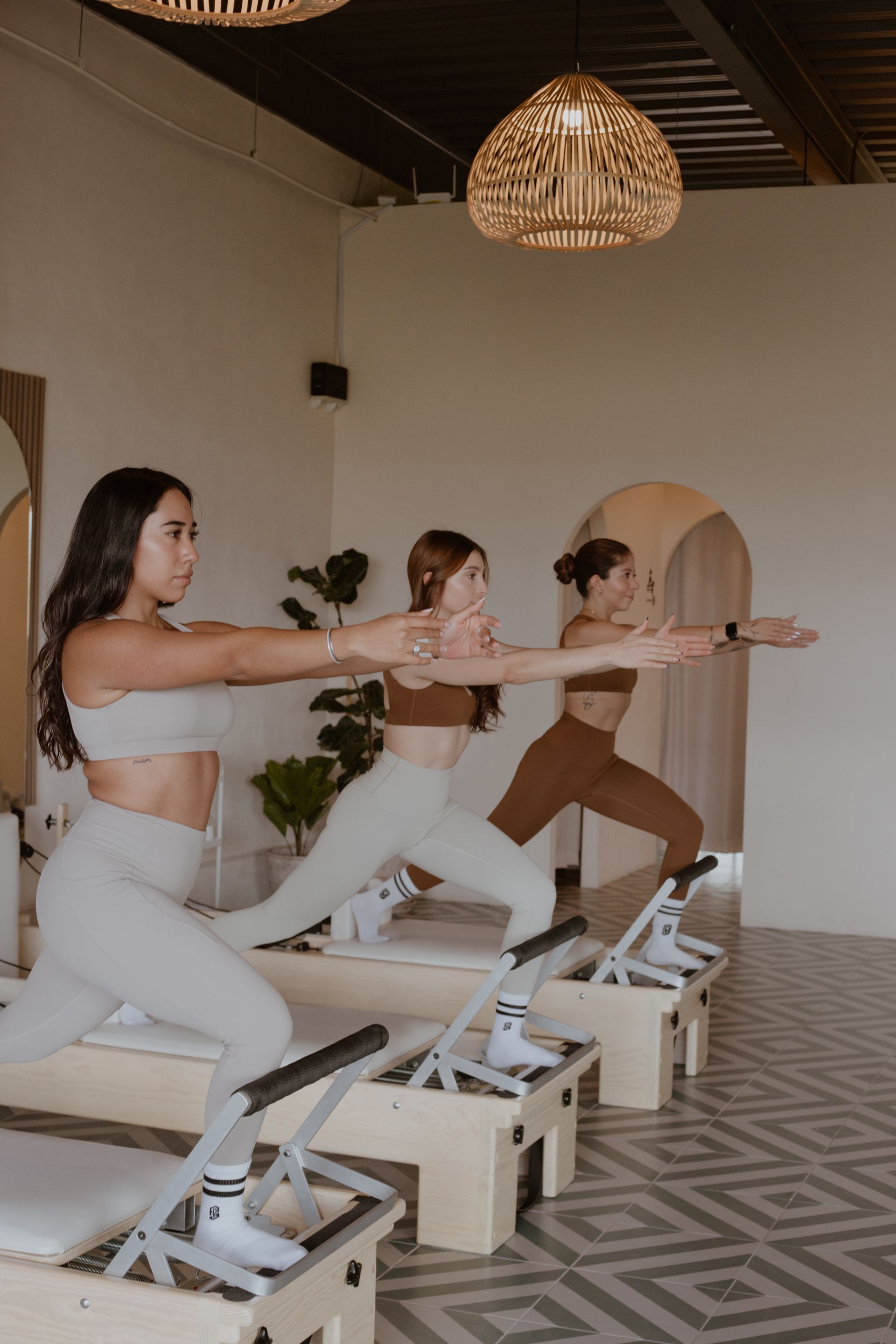 Tres personas practicando Pilates con reformer en un estudio luminoso con paredes blancas y lámparas colgantes tejidas.