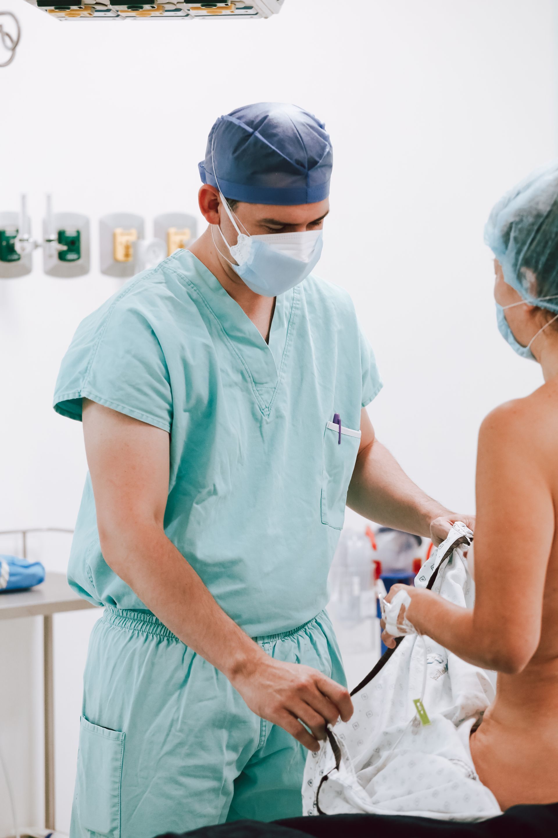 ROM&ROM  Médico con bata verde y mascarilla examinando a un paciente en una sala de exploración de una clínica médica.