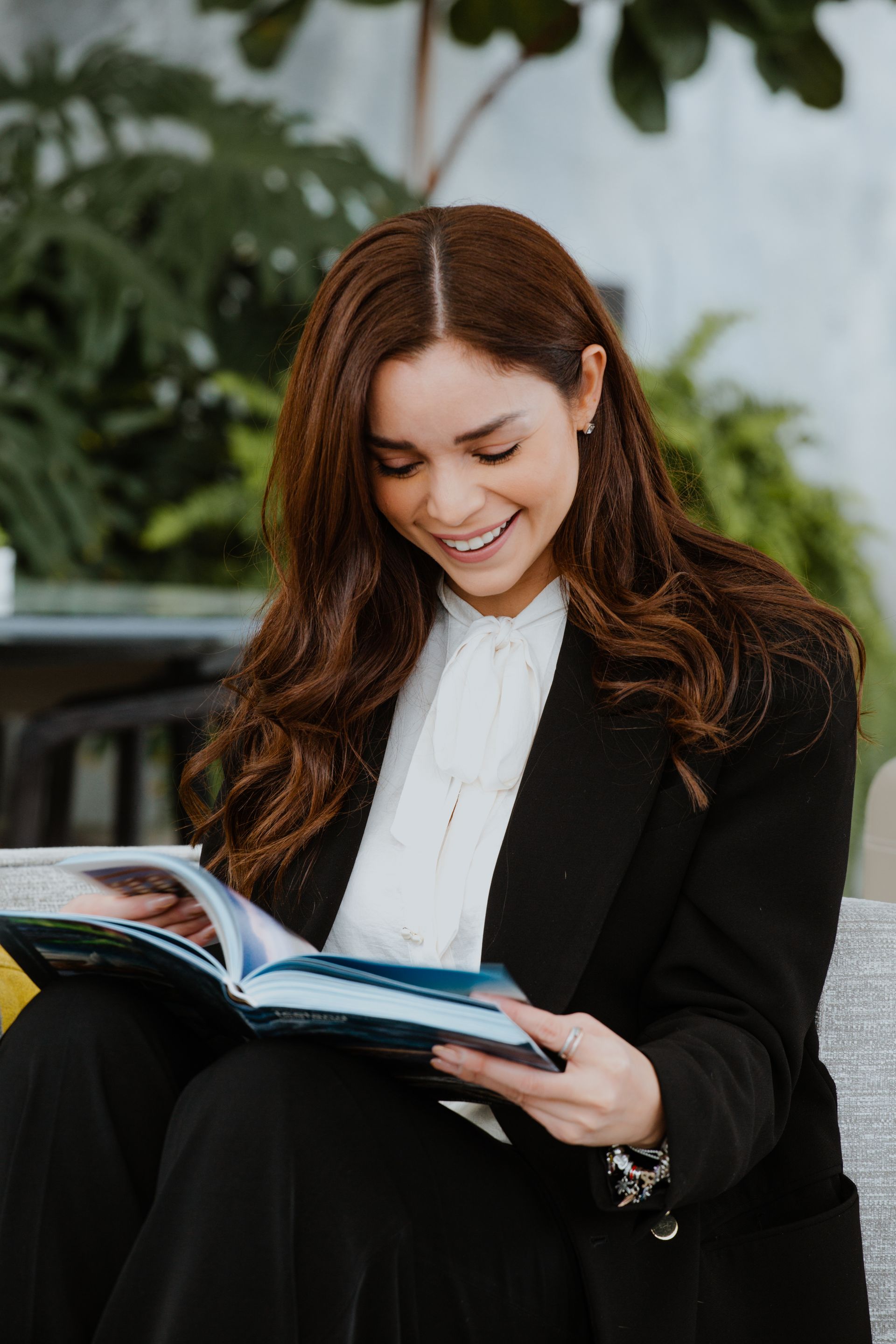 Mujer con blazer negro sonriendo mientras lee un libro en un luminoso ambiente interior.
