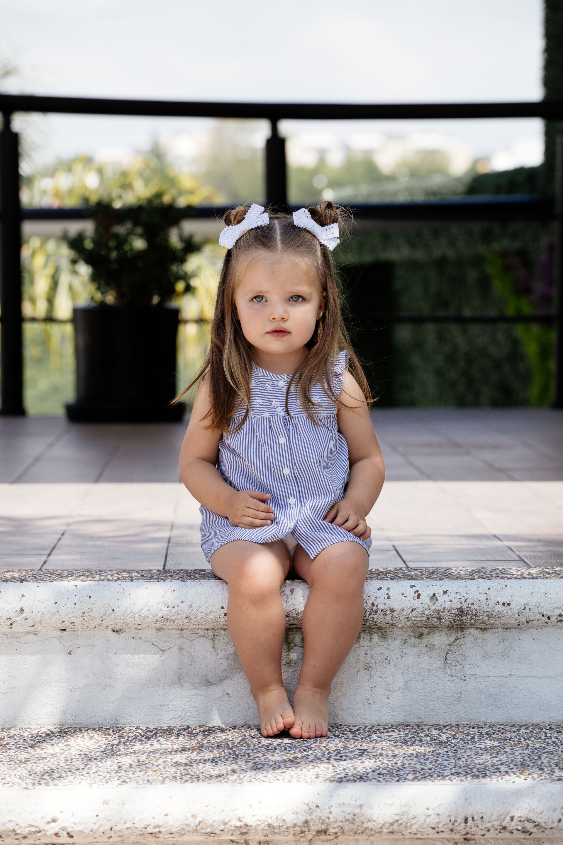 Una niña pequeña con un vestido de rayas está sentada en unas escaleras exteriores, mirando a la cámara.