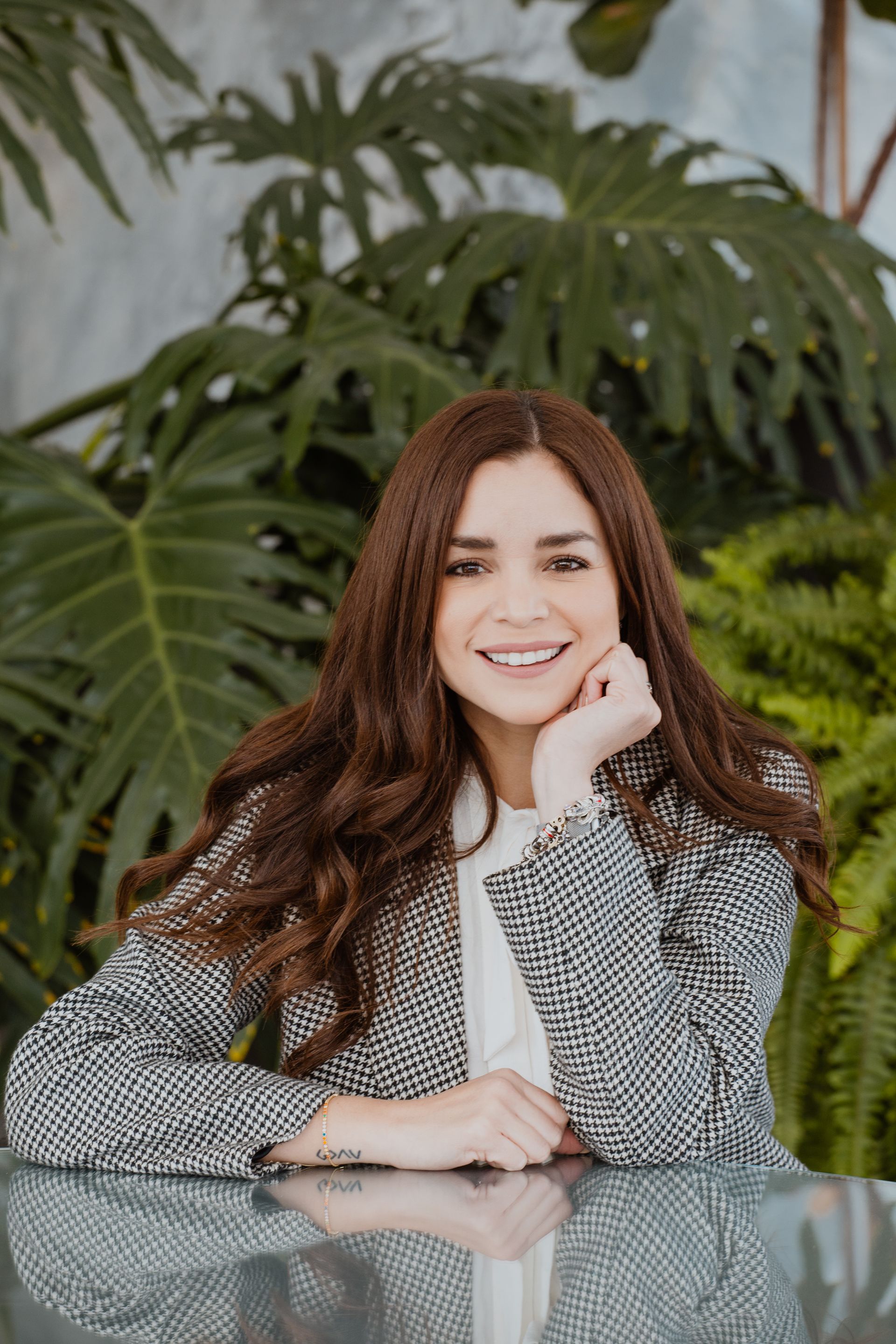 Mujer sonriente con chaqueta estampada apoyada en una mesa de cristal con exuberantes plantas verdes al fondo.