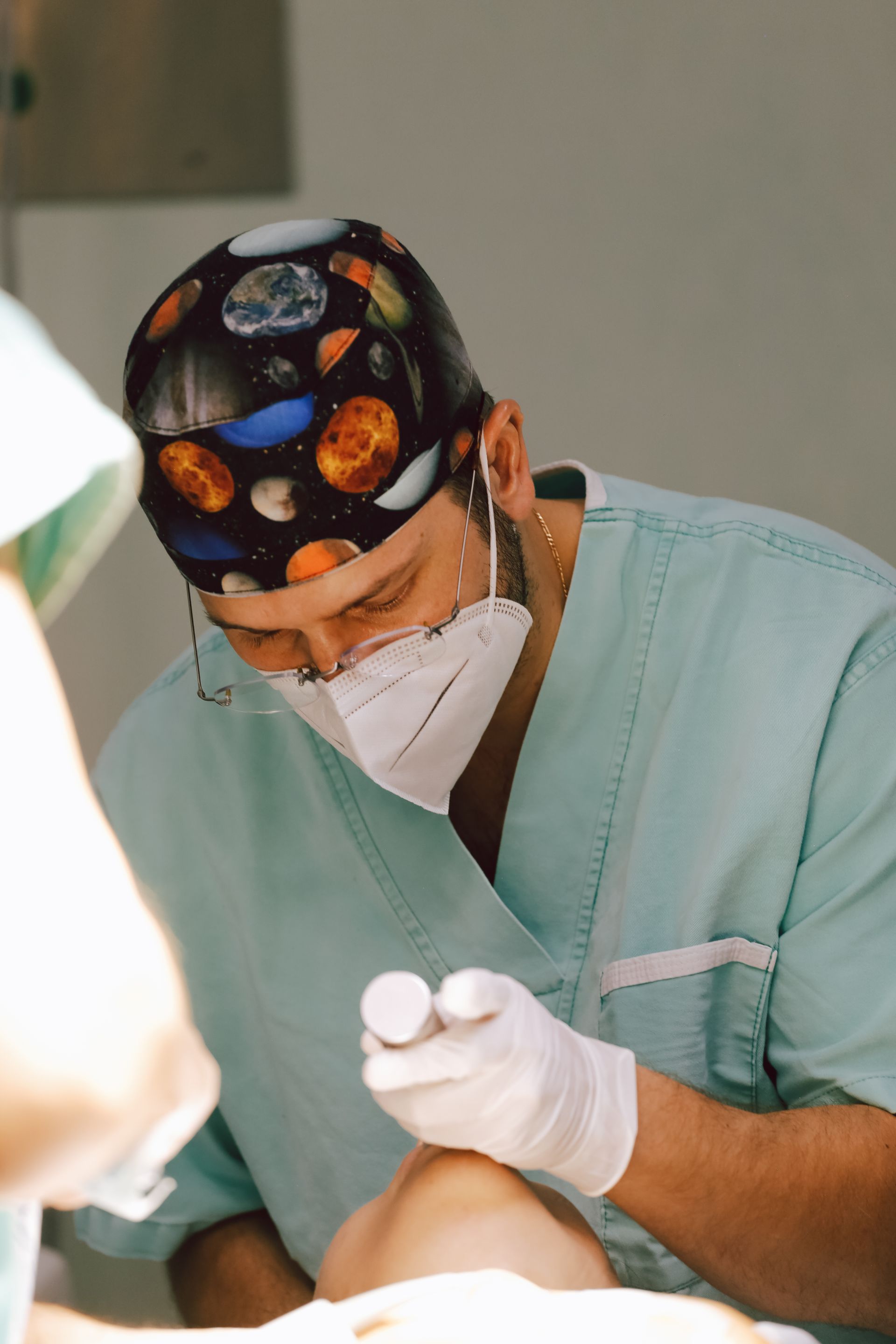 Cirujano con bata y mascarilla de color turquesa realizando un procedimiento médico con las manos enguantadas.