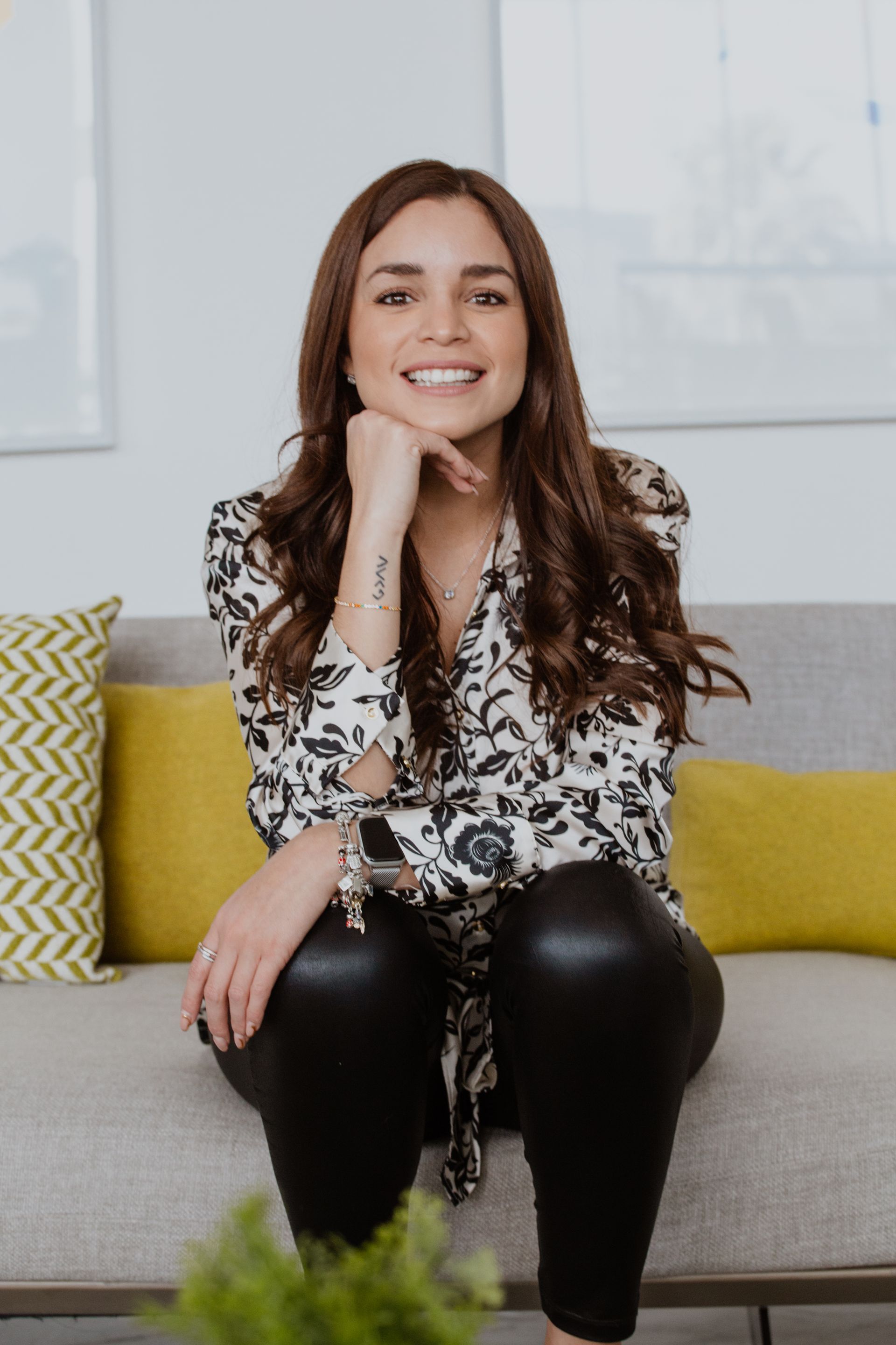 Mujer sonriente sentada en un sofá gris con cojines amarillos, vestida con una blusa blanca y negra y pantalones negros.