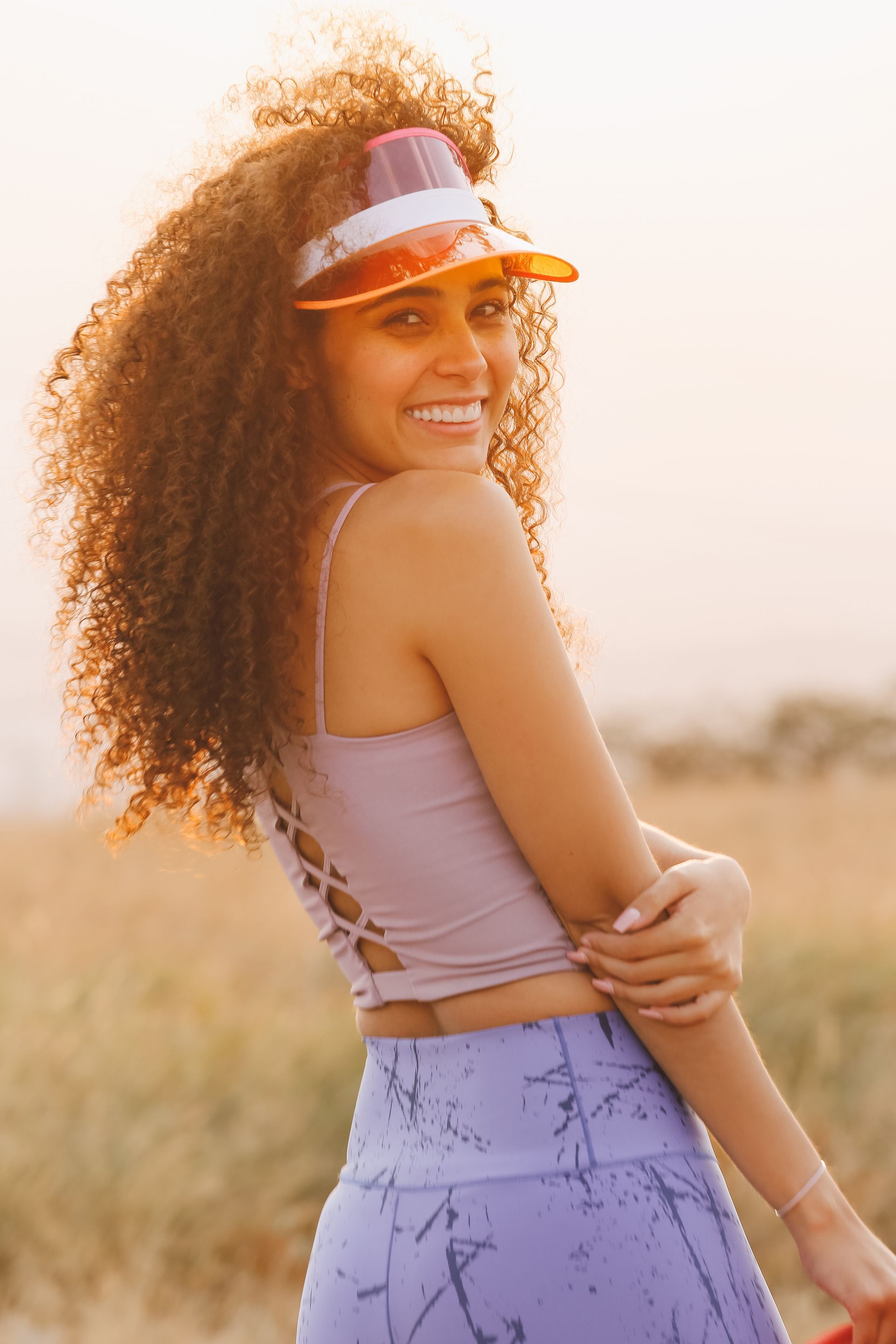 Mujer sonriente con visera blanca y atuendo lila de pie al aire libre al atardecer.